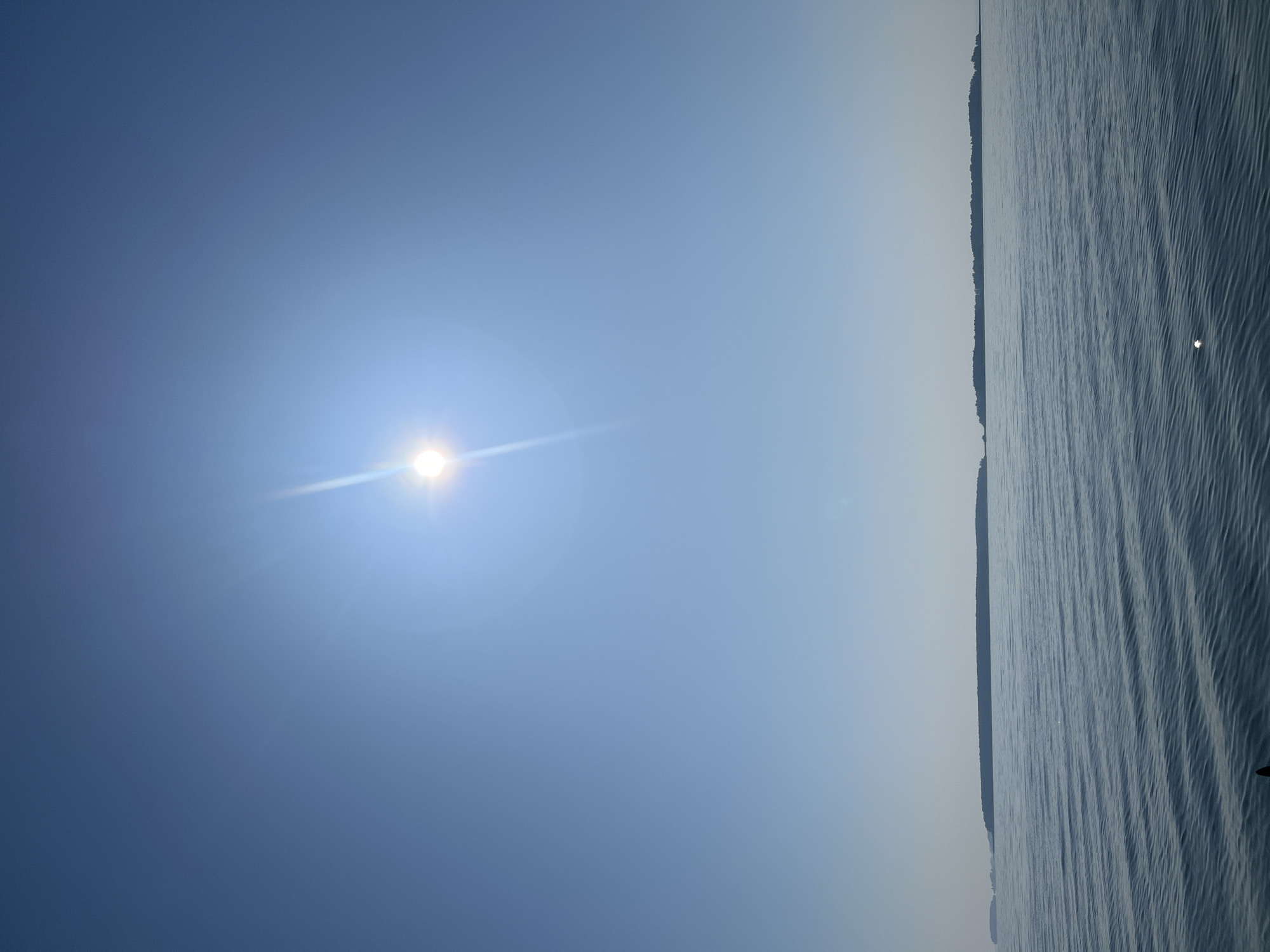 A vertical shot captures a serene, sun-drenched scene of a vast body of water under a clear blue sky. The sun, a brilliant orb with visible rays, dominates the upper portion of the image, casting a bright glare. The water below is calm, with gentle ripples reflecting the sunlight, creating a shimmering effect. In the distance, a long, dark silhouette of islands or a coastline stretches horizontally across the frame, creating a stark contrast against the bright sky and water. The overall impression is one of tranquility and expansive natural beauty. The location is the Ob River in Novopichugovo, Russia.