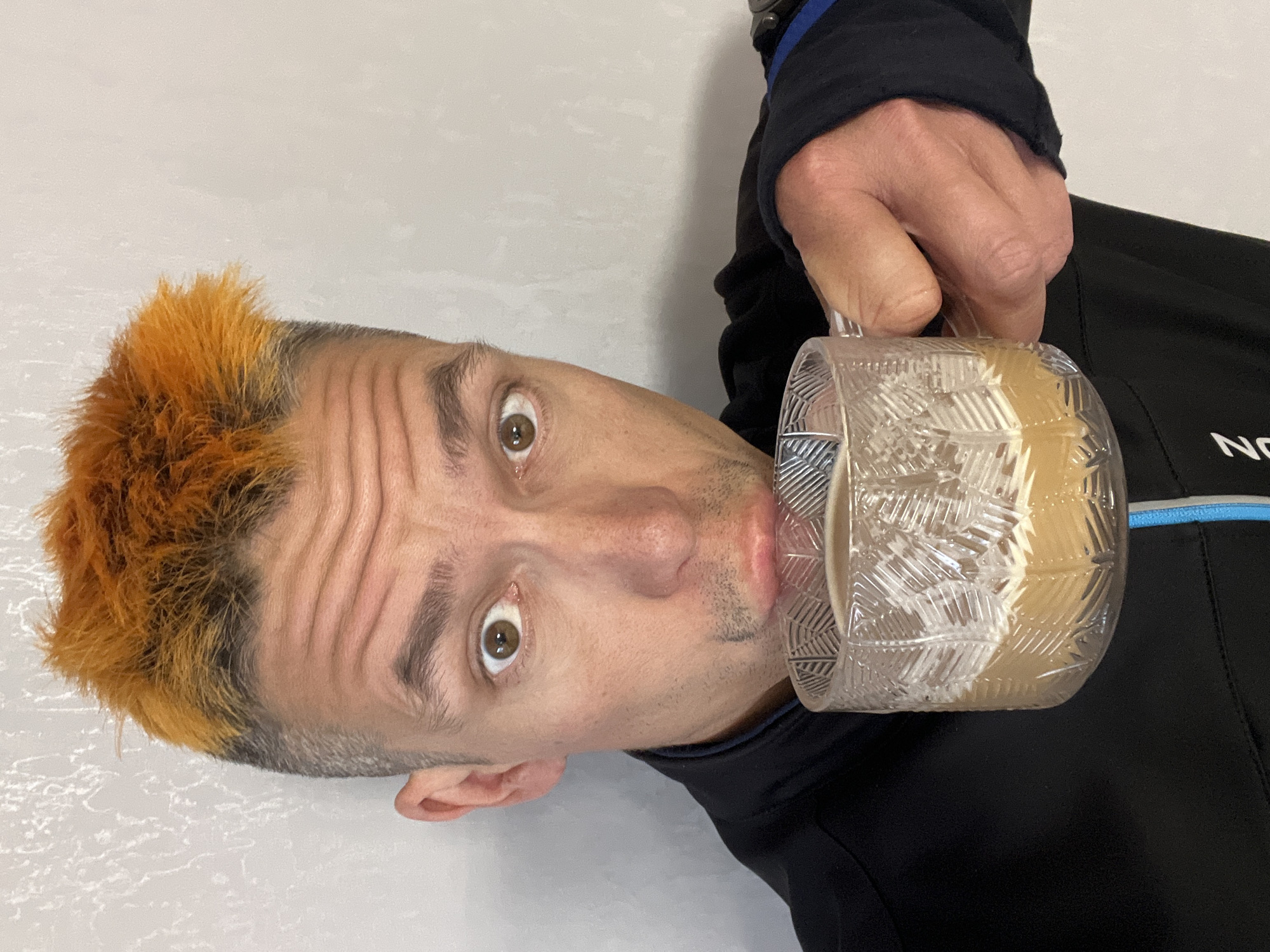 A person with graying hair and a bright orange fringe is drinking from a clear, patterned glass mug. The person is wearing a black jacket with a blue trim on the sleeve. The background appears to be a light-colored surface. The individual has light skin, brown eyes, and a short beard. No information is available to determine the specific location in Chaikovsky, Russia or any other identifying details.