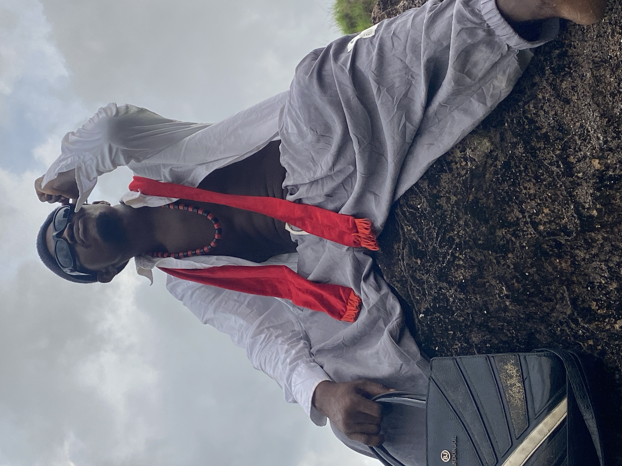 The image, taken in Nguzu Edda, Nigeria, features a dark-skinned male positioned outdoors against a cloudy sky. He is leaning or lying horizontally on a dark, rough, textured surface, which appears to be a rock formation.
The man is dressed in a white long-sleeved shirt, unbuttoned at the front, revealing a red and black beaded necklace. A bright red scarf or sash, with fringed ends, is draped around his neck and chest. He wears loose-fitting grey trousers. His head is covered by a dark beanie or cap, and he wears black sunglasses.
His right hand holds a black shoulder bag with a distinct design, featuring geometric ridges and a metallic diagonal stripe. Visible text on the bag includes "LONDON JOE" and a circular "BJ" logo. The background is composed solely of a grey, overcast sky, indicating a cloudy daytime setting. The individual appears to be in a relaxed pose, possibly resting or posing for the camera in a natural environment.