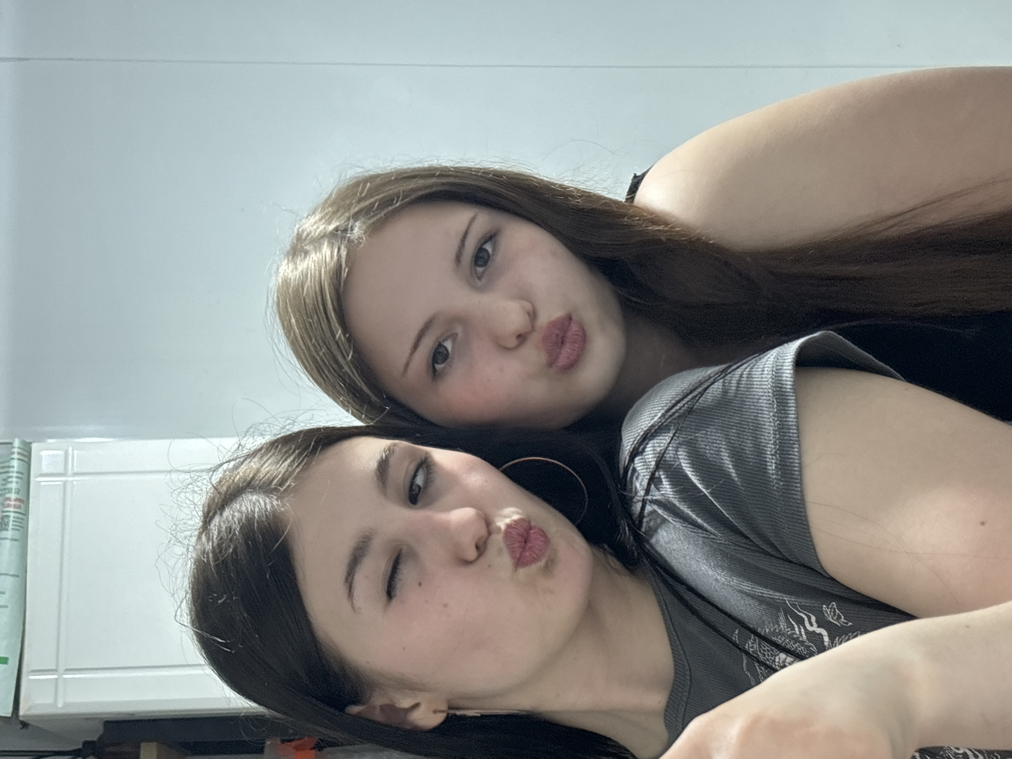 Two young women are posing closely together, likely indoors. The woman in the foreground is winking and puckering her lips in a kissing face. She is wearing a grey patterned top and a large hoop earring. The woman behind her is also puckering her lips. She has brown hair and is wearing a dark-colored top. The background is a white wall with some visible furniture or appliance. There is no indication of the location being in Фоки, Russia.