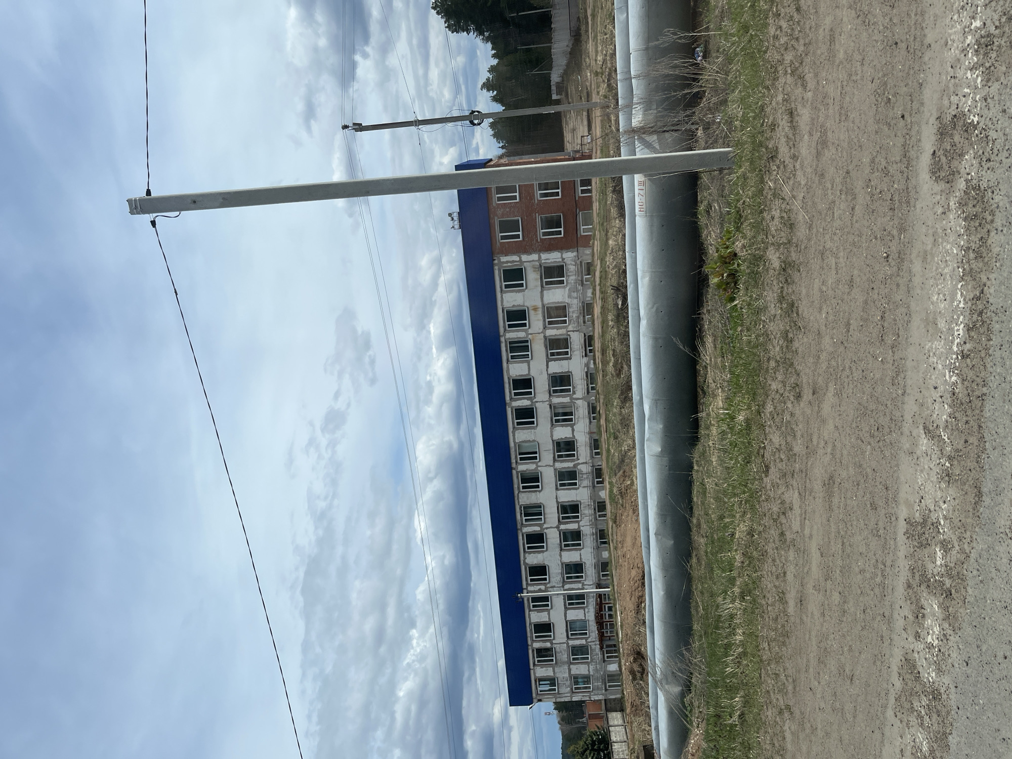 The image depicts a semi-industrial or residential area under a cloudy sky. The perspective is slightly elevated, looking towards a multi-story building and a dirt road.
In the mid-ground, a long, rectangular, multi-story building is visible, likely residential or commercial. It features a prominent blue roof and numerous windows, many appearing to be recent installations or part of an ongoing construction/renovation, set into light grey concrete block walls. Some sections of the building also show exposed red brick.
To the right of the building, and extending into the foreground, run several large, grey pipes, possibly for utilities like heating or water. These pipes are laid parallel to a dirt road or track that occupies the right side of the frame. The road surface is uneven, a mix of dirt and gravel, with a faint, broken white line visible along its right edge, suggesting it might be an old, unmaintained path. Sparse green grass and dry vegetation grow alongside the pipes and road.
Several utility poles with electrical wires crisscross the scene, some extending across the sky, others closer to the building and pipes. In the background, behind the building, a line of coniferous trees is visible, indicating a wooded area nearby. The sky is overcast with varying shades of grey clouds, suggesting daytime weather.
No people or active events are captured in the static scene. Visible text on one of the large grey pipes reads "Ш1 7-04 Ш". The setting appears to be an unadorned, utilitarian environment in Прикамский, Russia.