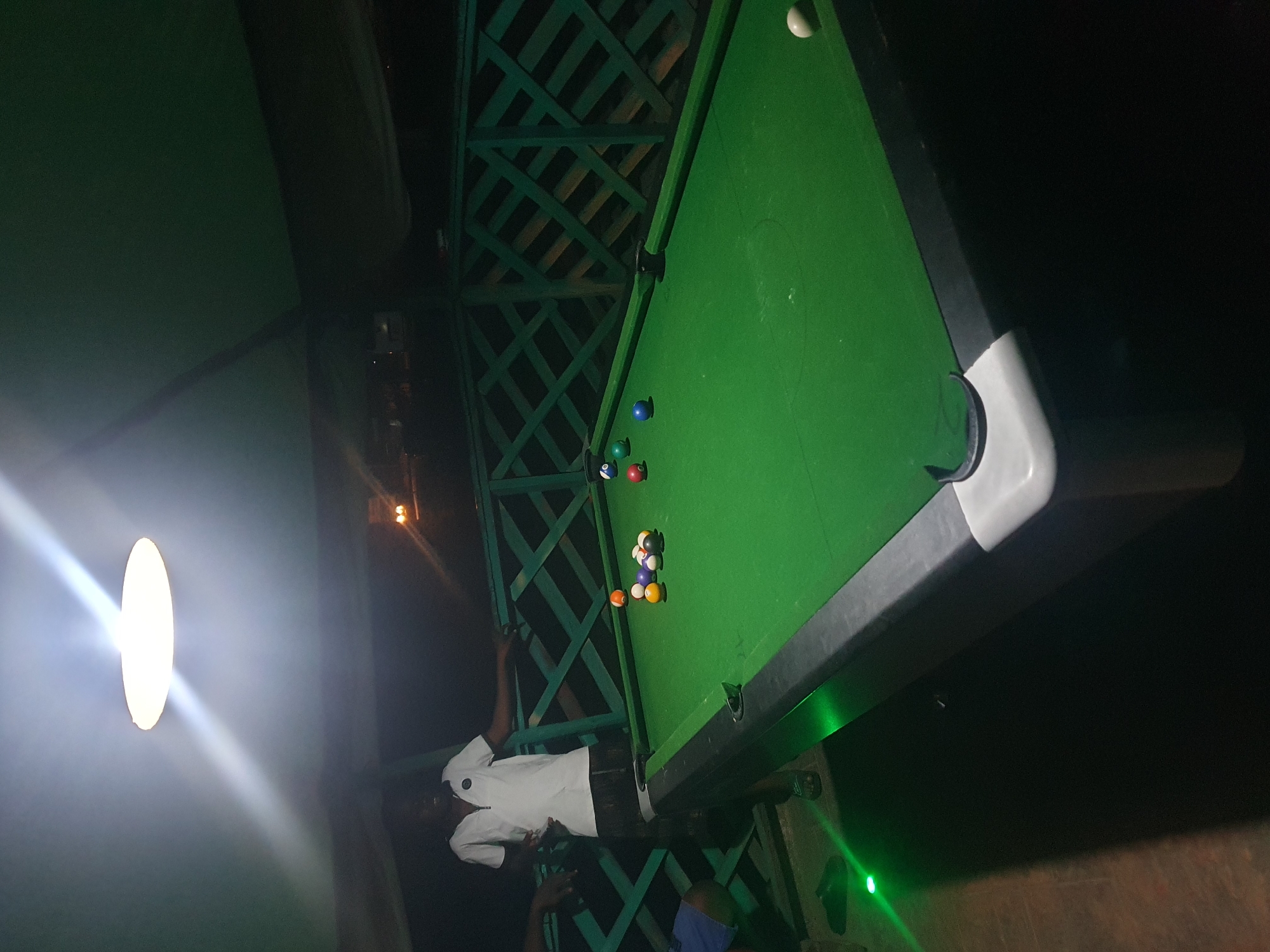 The image shows a pool table on an outdoor patio at night. A young man in a white polo shirt and dark shorts stands behind the table, leaning on a green railing. Several pool balls are scattered on the green felt surface of the table. The patio is lit by an overhead light fixture, and a green light shines on the floor near the man's feet. The background shows a dark night sky and a latticed green fence. The location is identified as Lokoja, Nigeria.