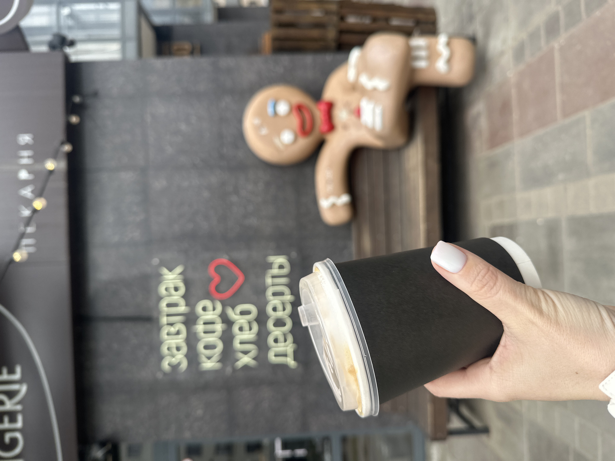 A hand holds a black disposable cup with a clear lid in front of a bakery sign in Krasnoyarsk, Russia. The sign lists "завтрак кофе хлеб десерты" (breakfast, coffee, bread, desserts) with a heart symbol. A gingerbread man figure sits on a bench in the background. The setting appears to be an outdoor area near the bakery.