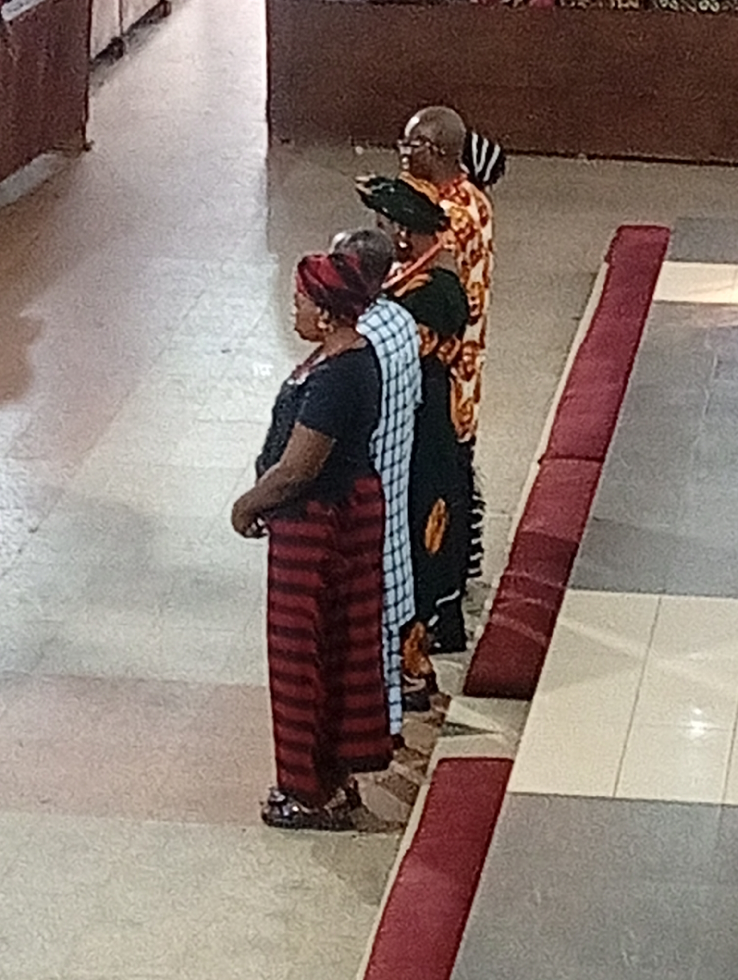 Certainly, here's a detailed description of the image:
**Scene:**
The image shows four people standing in a tiled hallway. A red carpet-like section runs along one side of the hallway.
**People:**
* **Person 1:** Closest to the viewer. An adult female with a red and black striped head wrap. She is wearing a black top and red and black striped pants.
* **Person 2:** To the right of Person 1, with a blue and white patterned dress or outfit, and a black head wrap.
* **Person 3:** Behind Person 2, wearing a black head wrap and a dark outfit with a gold design.
* **Person 4:** Farthest back, wearing a patterned orange and white top, glasses, and a striped hat or head wrap.
**Location:**
The location appears to be indoors, possibly in a building with tiled floors and neutral-colored walls. The lack of any additional identifiers in the request prevents determining the city of Unknown, Unknown.
**Objects:**
* Tile floor
* Red carpet section
* Head wraps
* Clothing
**Actions/Interactions:**
All individuals appear to be standing still, possibly observing something out of view of the camera. There is no discernible interaction between them.