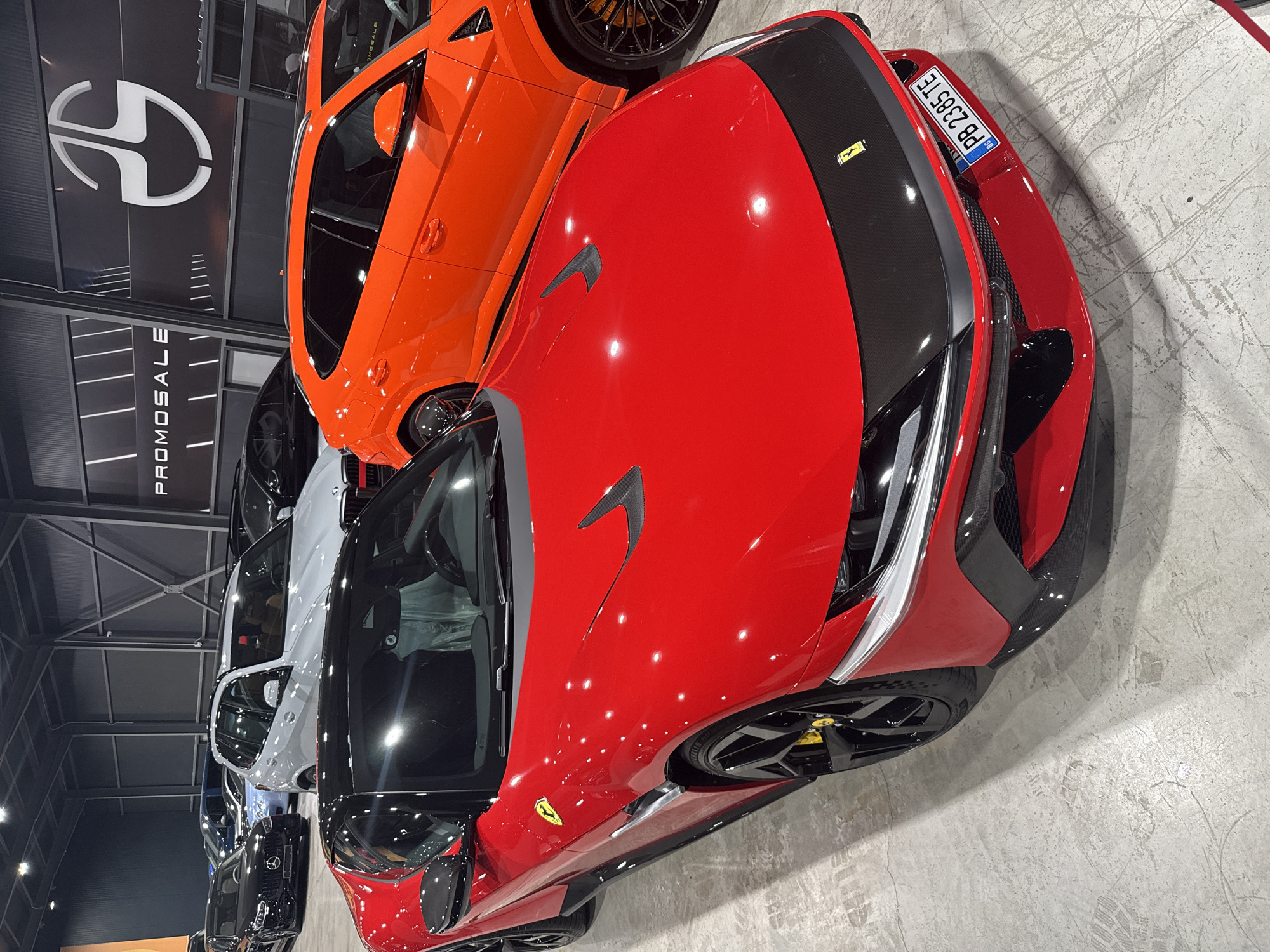 The image shows the interior of a car dealership, possibly located in Plovdiv, Bulgaria, as indicated by the license plate on the red Ferrari which reads "PB 2385 TE", a Bulgarian format. In the foreground, a bright red Ferrari is parked, facing left, with its front end prominently displayed. The car features a black hood accent and a black front splitter, with a yellow Ferrari emblem on the fender. The license plate is clearly visible. Behind the Ferrari, an orange Lamborghini SUV is parked, partially obscured. Further in the background, several other vehicles are visible, including a white BMW SUV and a black sedan. The dealership has a high ceiling with industrial lighting. A large logo, possibly for "PROMOSALE," is visible on the back wall. There are no people visible in the image.