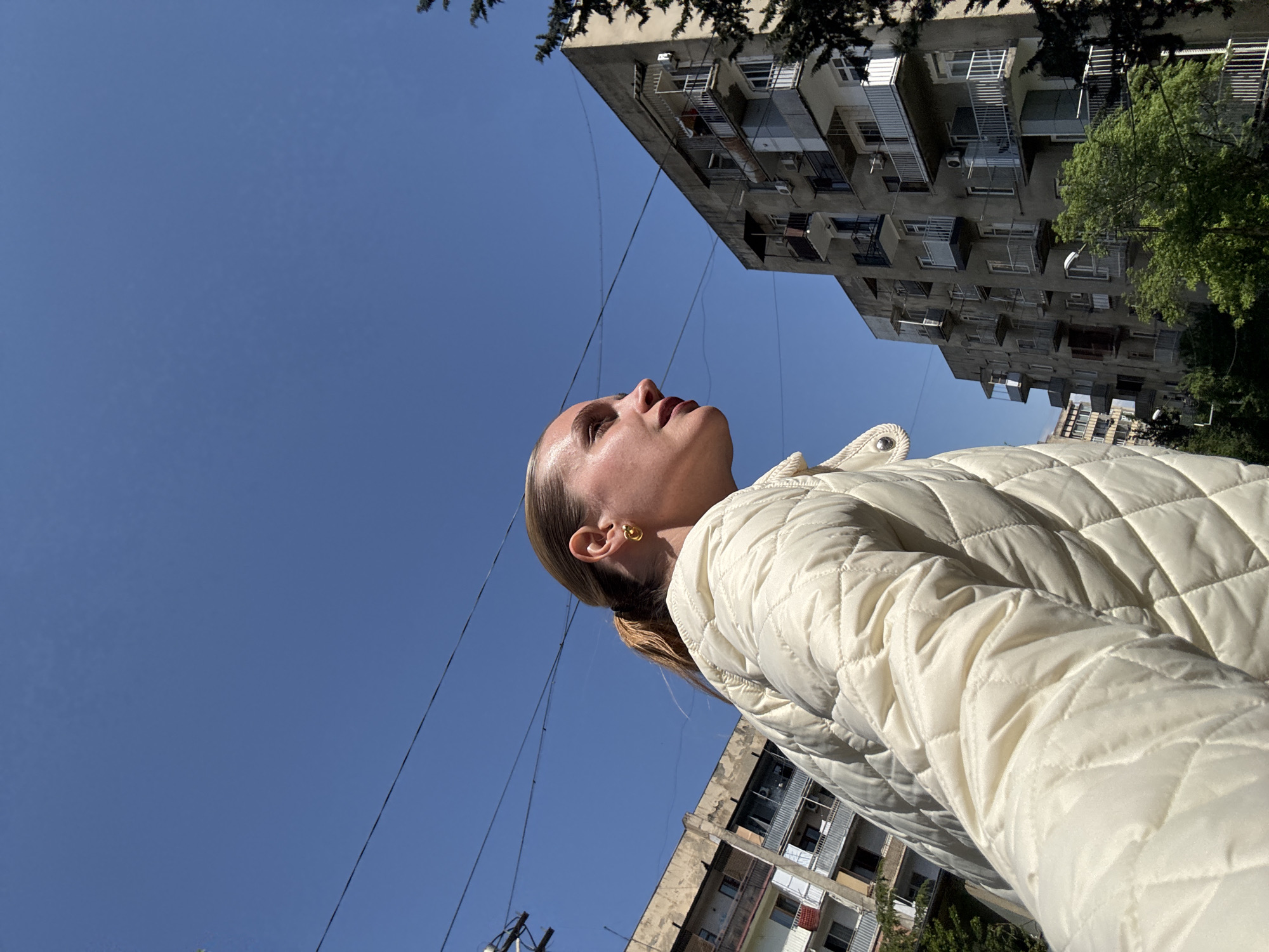 The original image, rotated 90 degrees clockwise, offers a low-angle perspective of an urban setting. The main subject is a woman, positioned on the right side of the frame, looking upwards and to the left. Her profile is clearly visible, with her hair pulled back and a gold-toned earring on her ear. She wears a light, possibly cream-colored, quilted jacket with a visible button on the collar. Her expression is neutral and observant as she gazes into the bright, clear blue sky that occupies a significant portion of the image, particularly to the left and top.
In the background, several multi-story concrete apartment buildings rise prominently. These structures feature numerous balconies, some enclosed, and display a characteristic post-Soviet architectural style, consistent with the location context of Tbilisi, Georgia. Green foliage from trees is visible on the far left and top right edges, interspersed among the buildings. Thin black utility wires crisscross against the vivid sky, adding to the urban landscape. The scene is illuminated by bright daylight, suggesting sunny weather during the day, creating distinct contrasts and highlights. There is no visible text in the image.