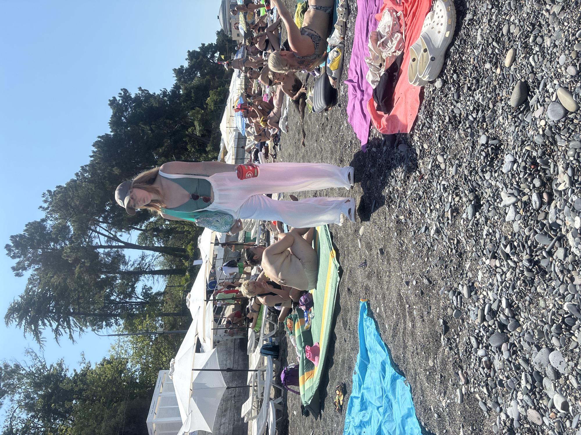 A woman wearing a green top and white pants, a baseball cap, and sunglasses stands on a pebble beach in Sochi, Russia. She is holding a blue bag and a red can. Many people are sunbathing on lounge chairs and on towels. There are white umbrellas providing shade. The water is not visible in the frame.