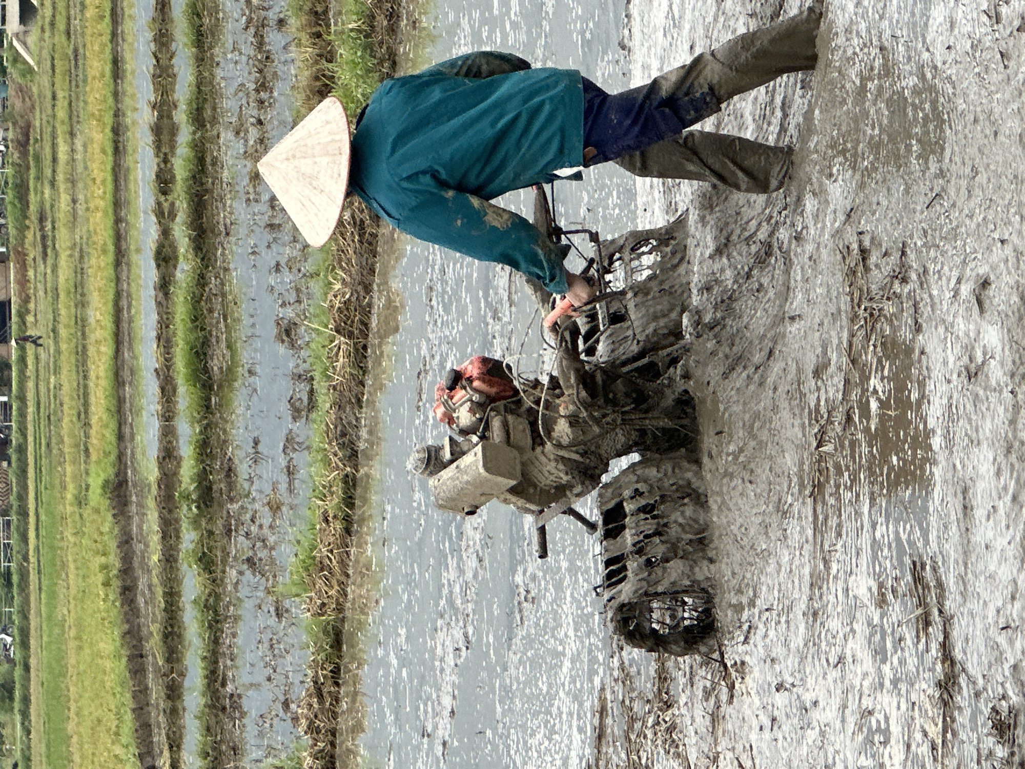 A person wearing a conical hat and a teal jacket is tilling a muddy rice paddy in Mai Chau, Vietnam. The person is pushing a tiller, which is partially submerged in mud and water. The rice paddy is surrounded by green fields and appears to be in the process of being prepared for planting.