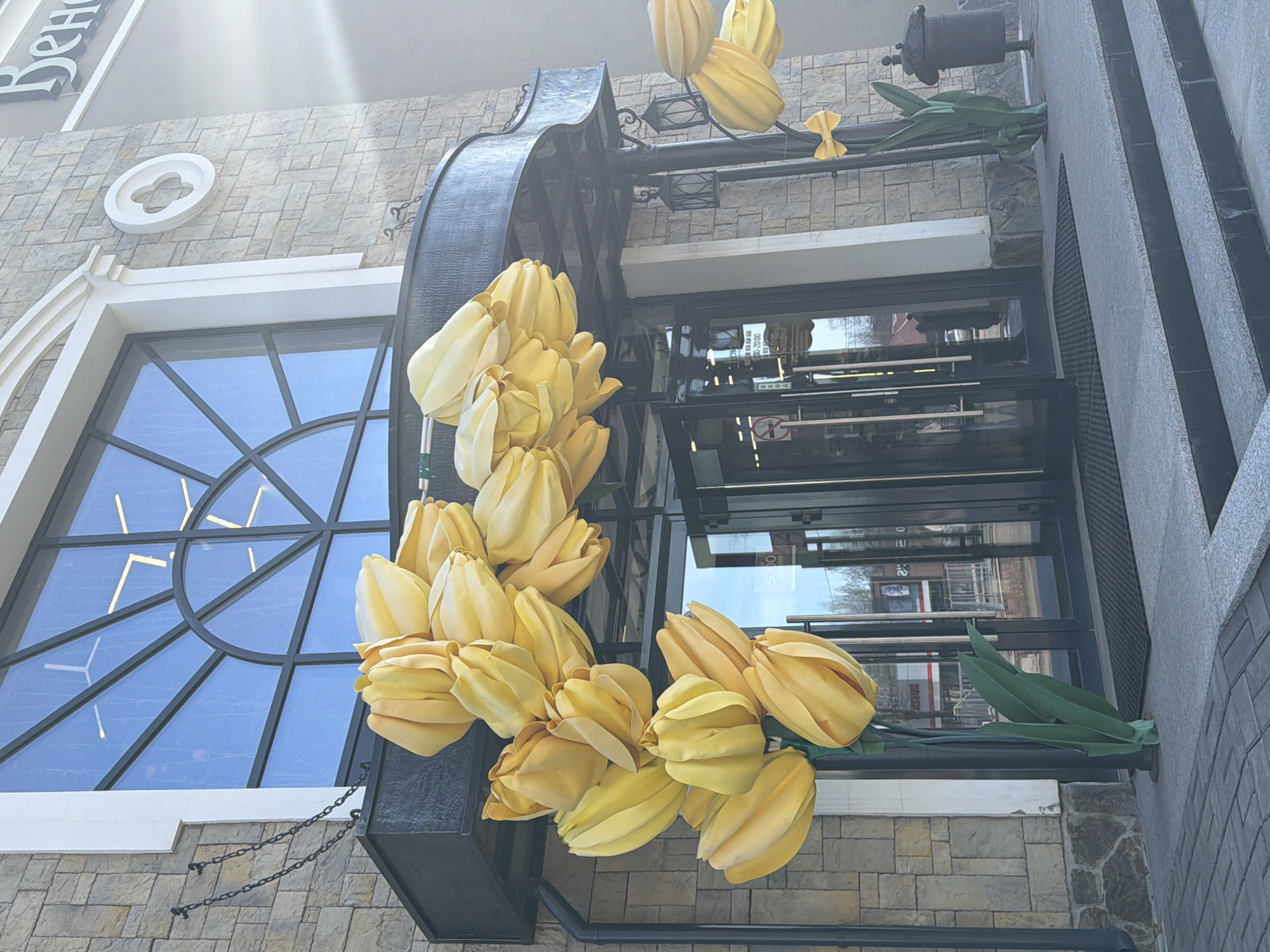 The image shows a building facade, possibly a storefront, with a large window and decorative elements. A black metal frame adorned with large yellow tulip-like flowers wraps around the building's entrance. The window has a geometric design. The building appears to be constructed with gray stone. A sign reading "Вено..." is visible on the upper left. The location is suspected to be in Votkinsk, Russia.