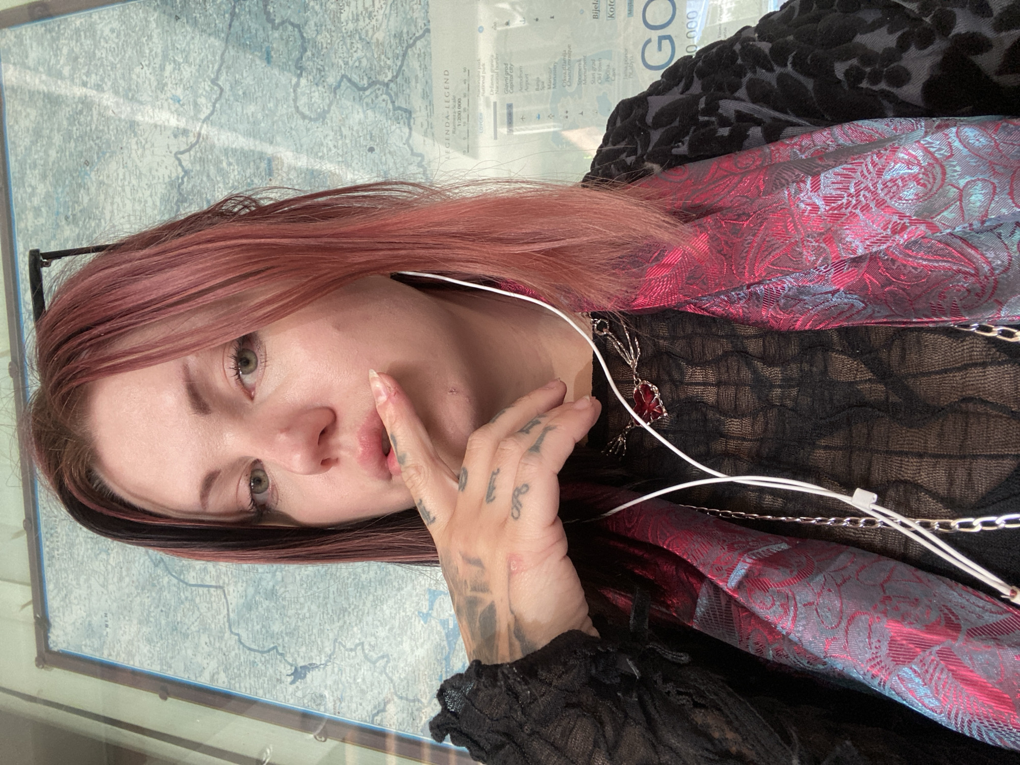 The image shows a person with pinkish-red hair, wearing black lace clothing and a red and black patterned shawl. They are wearing headphones and a necklace with a red pendant. Their hand is near their mouth, and tattoos are visible on their hand and arm. Behind the person is a framed map, titled "LEGENDA-LEGEND" and "Koto" at the top right, with text indicating it shows a scale, national parks, national borders, capital cities, airports, spas, monasteries, churches, mosques, monuments, old towns, and caves. "Budva, Montenegro" isn't present in the image.
