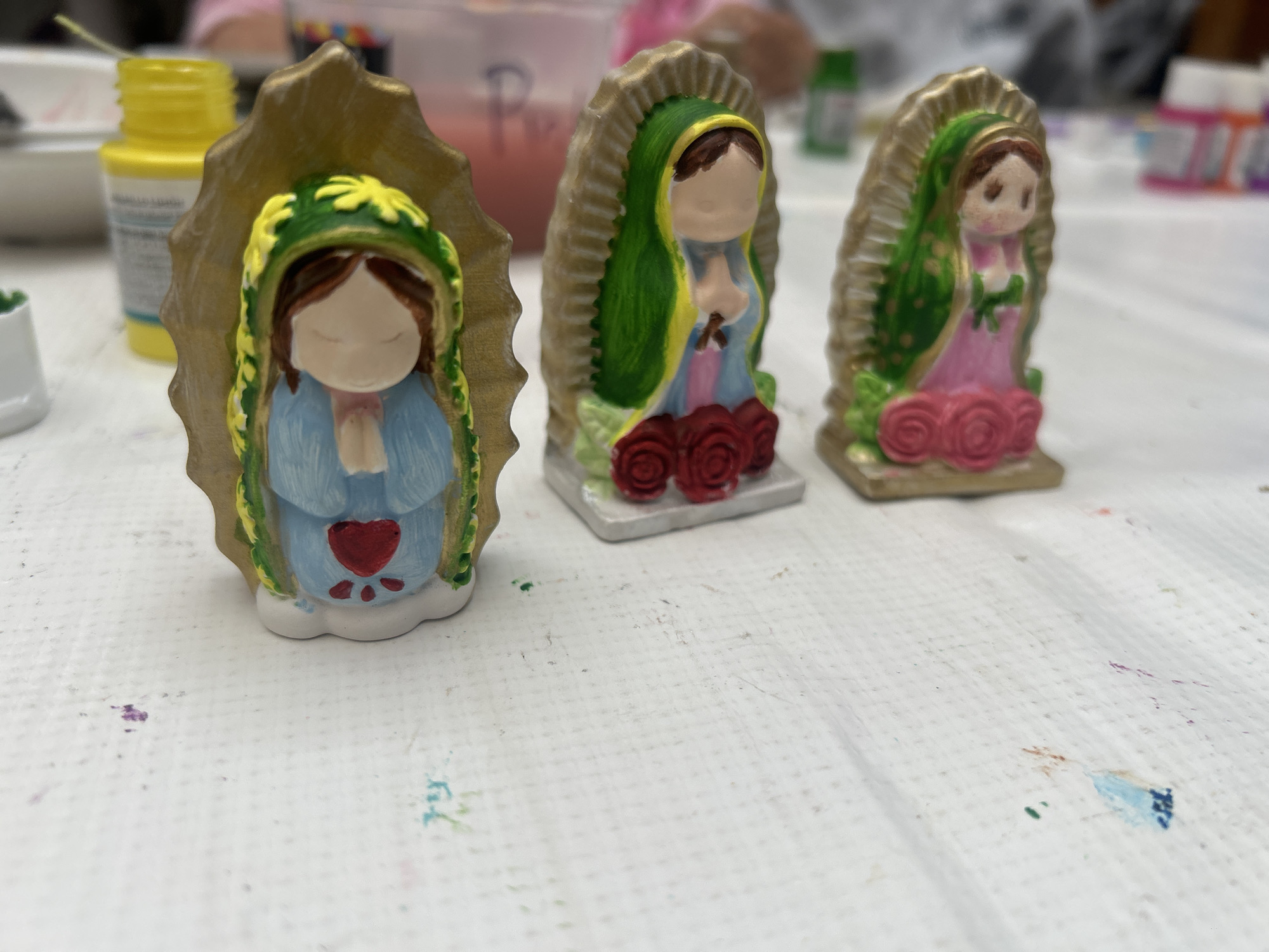 The image shows three small, painted figurines of the Virgin Mary standing on a white, textured surface. The figurines are positioned in a line, from left to right. Each is enclosed in a golden frame-like structure and appears to be hand-painted with different color schemes. The leftmost figurine is painted with a blue robe, green and yellow head covering, and has a red heart on the robe. The middle figurine has a green cloak, yellow details, and red roses at the base. The rightmost figurine features a green cloak and a pink robe with pink roses. In the background, there are blurred objects including containers of paint, suggesting an art or craft setting. The location is Unknown, Unknown.