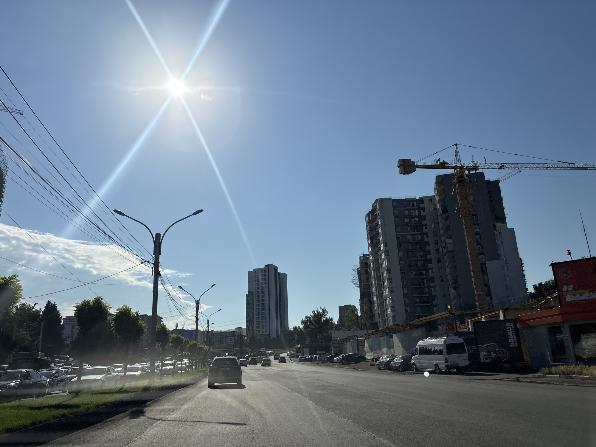 The image depicts a sunny day in Tbilisi, Georgia, with a view of a city street lined with trees and buildings. The sun is bright in the sky, causing lens flare. A construction crane is visible, along with several tall apartment buildings, some of which appear to be under construction. Traffic is present on the road, with various cars and a white van. Overhead power lines crisscross the sky. The scene suggests an urban environment with ongoing development.