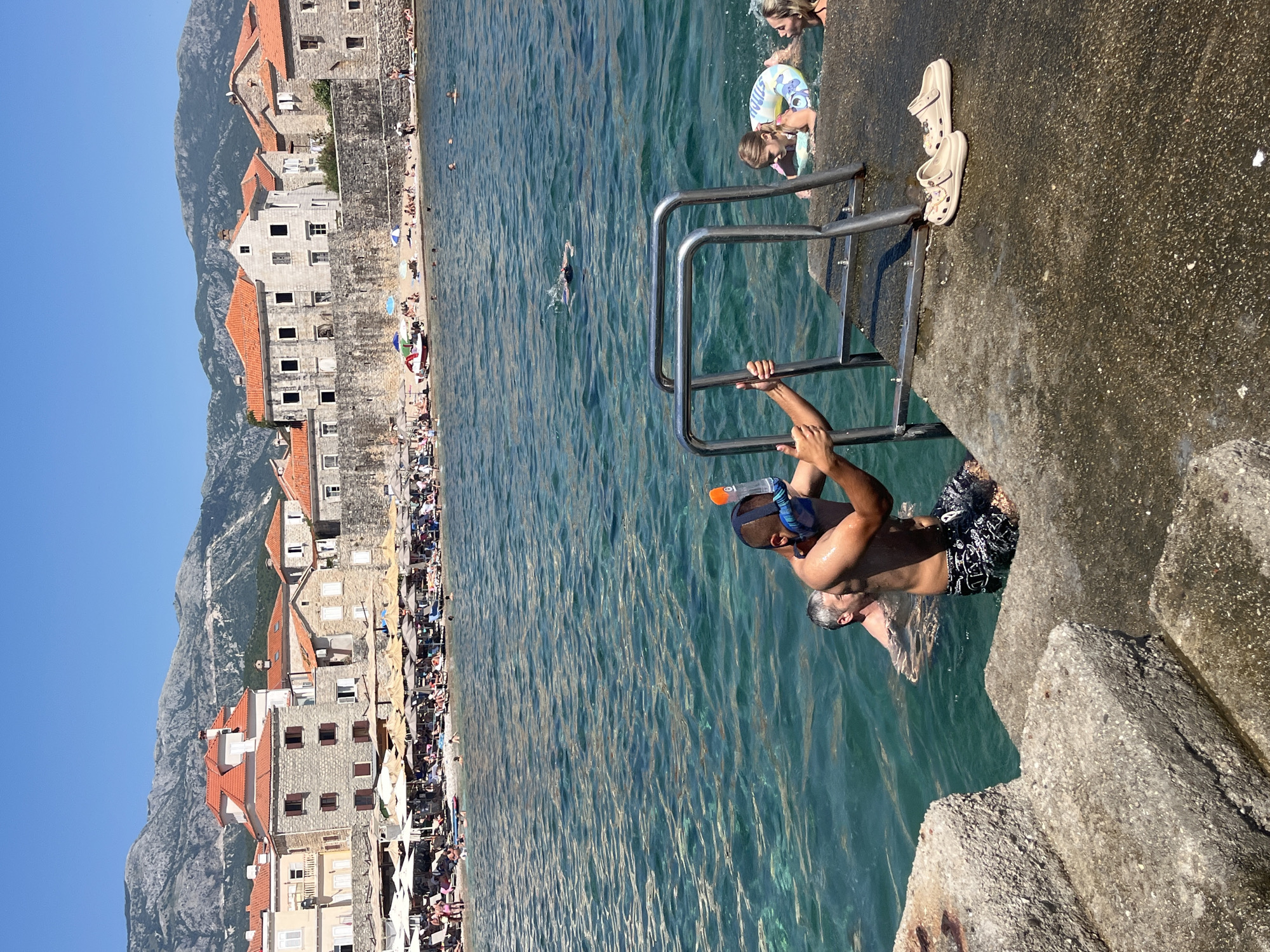 This media file captures a lively summer scene on a sunny day in Budva, Montenegro. The image is oriented vertically, showing a picturesque coastline with old stone buildings and red-tiled roofs leading up to a mountain backdrop. A crowded beach with numerous sun umbrellas and people relaxing is visible along the shore.
The foreground focuses on a concrete pier with a metal ladder descending into the clear, turquoise water. Two individuals are prominently featured. One person, wearing a snorkel mask and swim trunks, is holding onto the ladder and appears to be preparing to enter the water. Another person, partially submerged in the water, is visible nearby. In the background, several other people are swimming and enjoying the sea. A few more individuals, including children, are at the edge of the pier, with one child wearing a colorful inflatable ring.
The overall atmosphere is one of vacation and recreation, with a bright, sunny sky suggesting warm weather. The architecture of the buildings and the mountainous terrain are strong indicators of the Mediterranean location. The scene is bustling with activity, reflecting a typical summer day at a popular coastal resort. There is no visible text in the image.