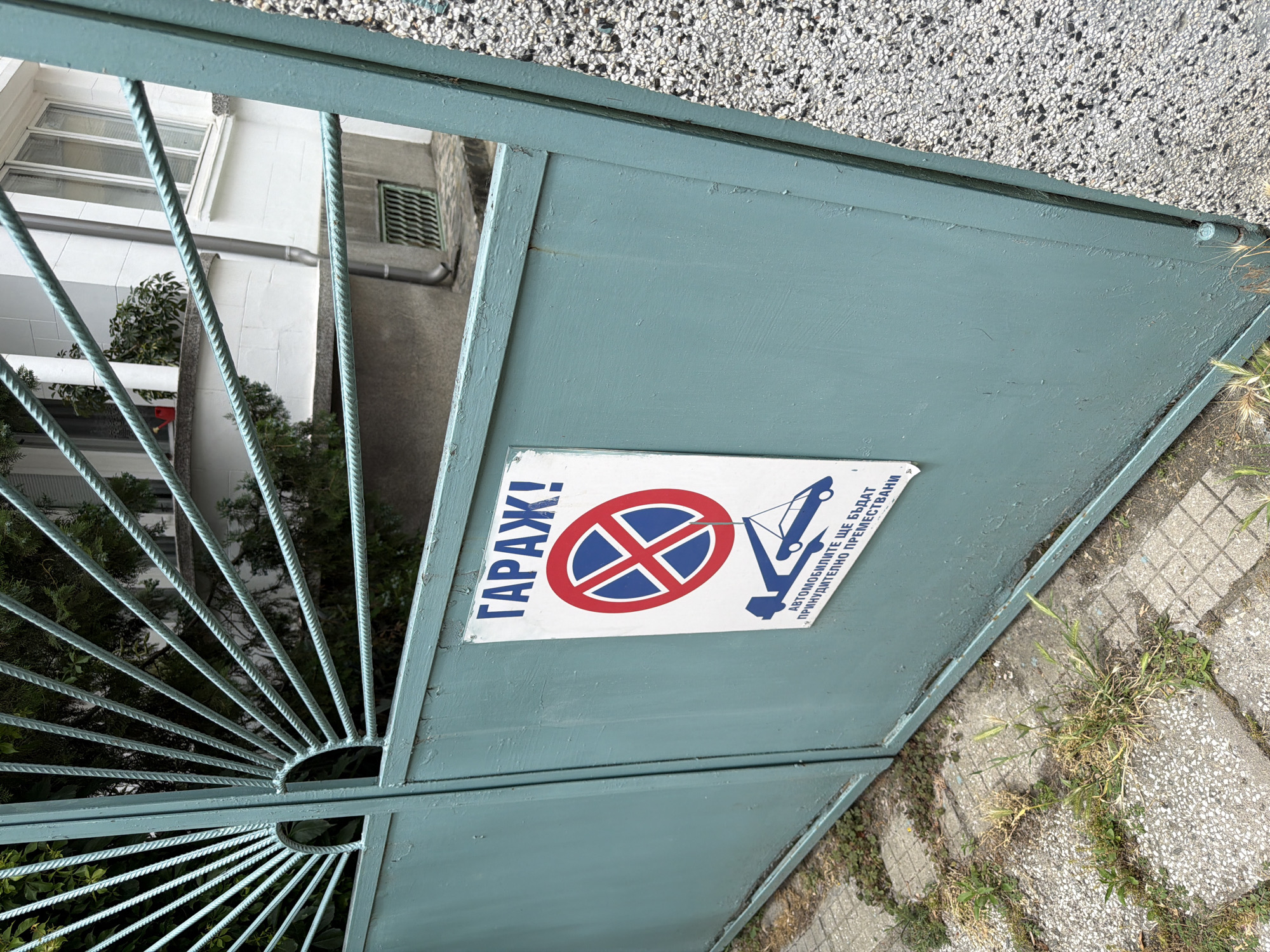 The image shows a close-up view of a turquoise metal gate with a sign attached. The sign has a red and blue circular prohibition symbol with a diagonal red line, and below it, a depiction of a tow truck lifting a car. Text on the sign, in Bulgarian, reads "ГАРАЖ!" (Garage!) at the top and "АВТОМОБИЛИТЕ ЩЕ БЪДАТ ПРИНУДИТЕЛНО ПРЕМЕСТЕНИ" (Cars will be forcibly relocated) below the graphic. The gate is closed. Behind the gate, some greenery is visible, and a white building with windows and a drainpipe can be seen. The ground in front of the gate is paved with concrete tiles, with some weeds growing between them. The environment suggests an outdoor urban setting.