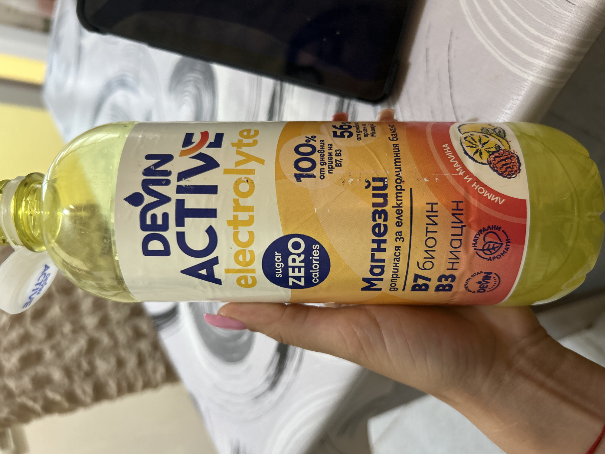A hand with a pink fingernail holds a clear plastic bottle of "DEVIN ACTIVE electrolyte" drink. The bottle's label is primarily white with blue, yellow, and red text. The drink inside is a pale yellow liquid. The background is blurred and appears to be a table with a patterned cloth. A portion of a black mobile phone is visible on the right side of the image. The lighting is somewhat diffused.