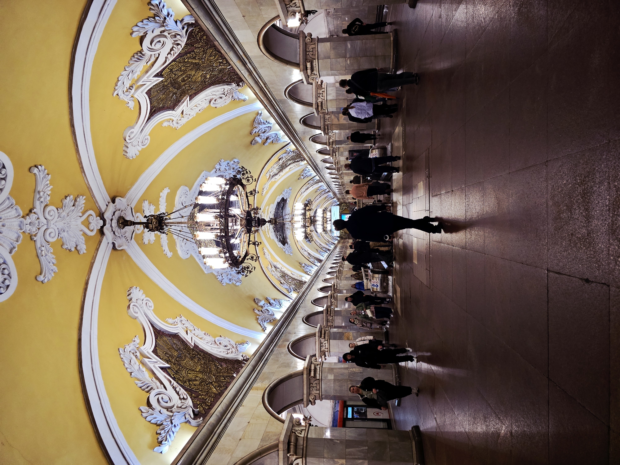 The image depicts a subway station in Moscow, Russia. The station features a long, arched ceiling painted yellow with white decorative molding and ornate chandeliers. Passengers are walking along a polished floor in the station, some standing near ticket machines. The station is well-lit and ornate.