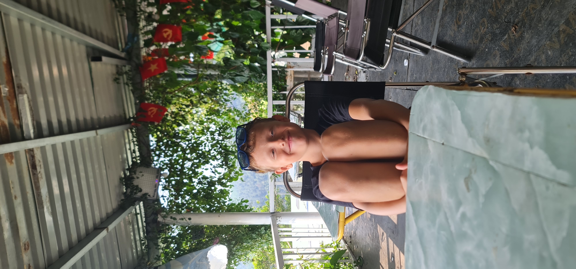 A young boy with sunglasses perched on his head sits at a table in an outdoor setting. He is fair-skinned with short blonde hair and is wearing a dark-colored t-shirt. His knees are drawn up towards his chest. The table is light green. Other chairs are visible behind him. The background includes trees, Vietnamese flags, and a white railing. The location is Đà Nẵng, Vietnam.