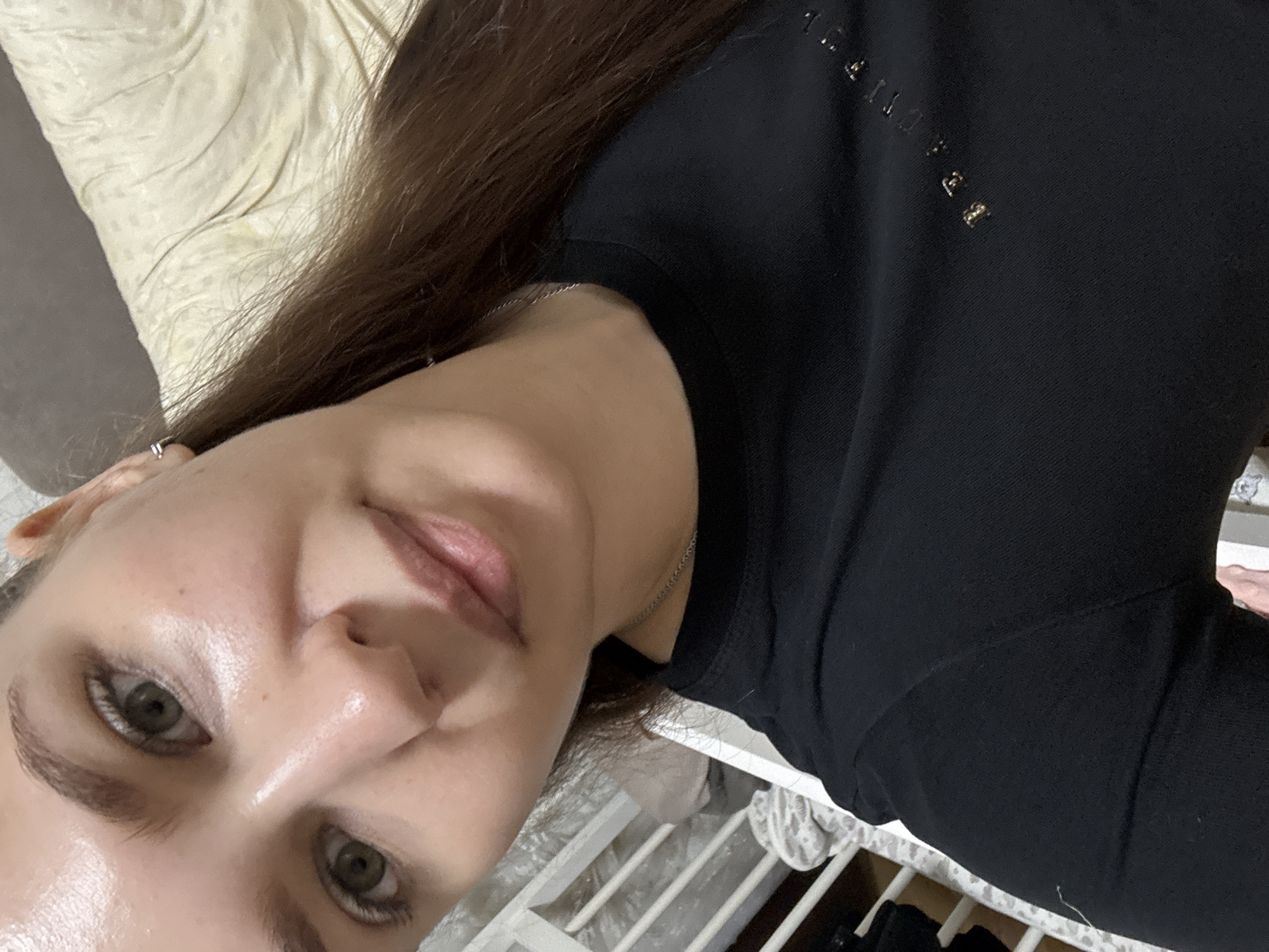 A woman with long brown hair is lying on a bed with a light-colored headboard. She is wearing a black shirt with some silver lettering on the upper right side. The background appears to be a bedroom setting. The location, Chaikovsky, Russia, cannot be determined from the image alone.
