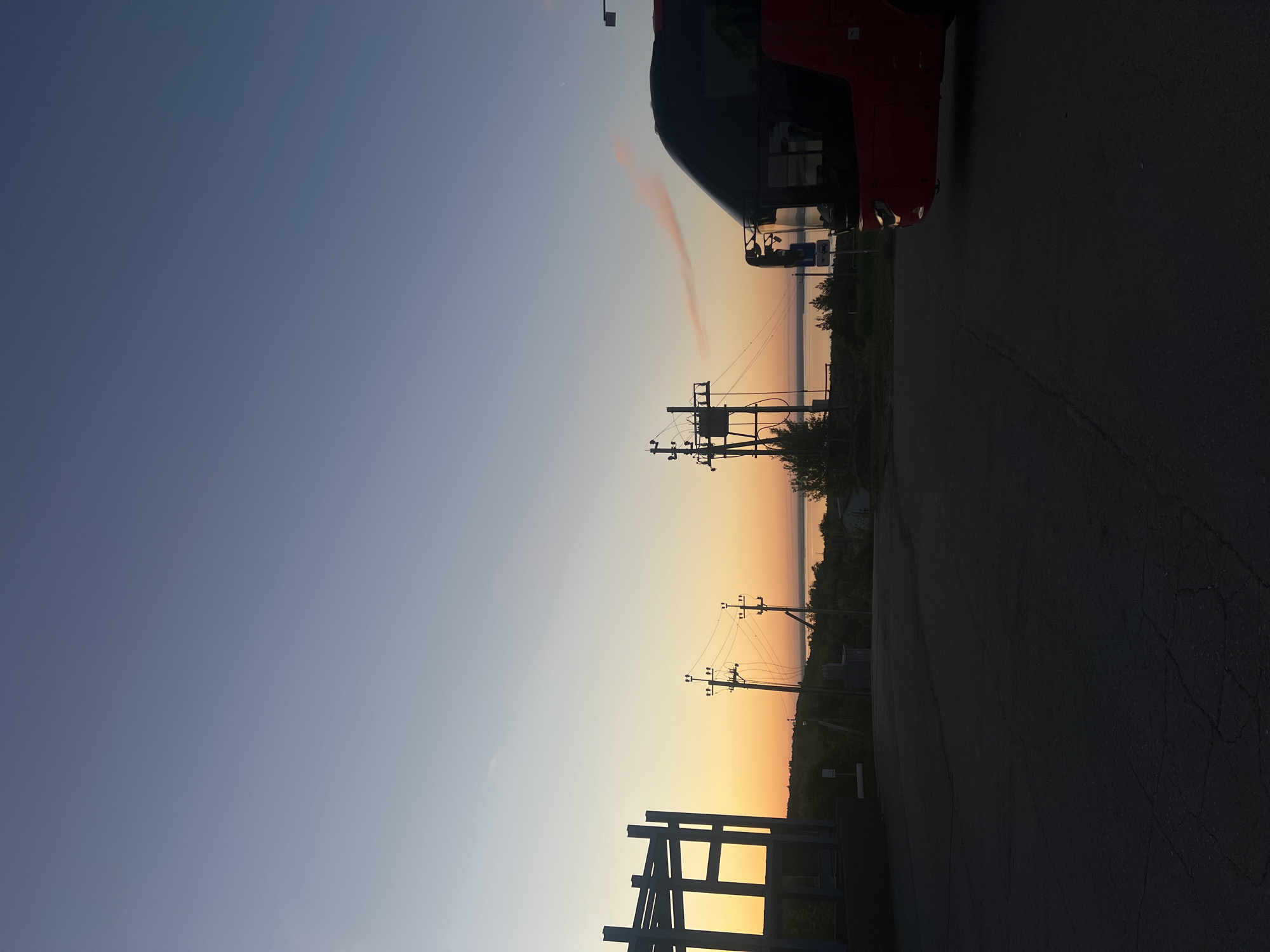 The image captures a sunset in Khvalynsk, Russia. Silhouetted power lines stretch across a horizon where the sky transitions from blue to orange. A road occupies the foreground with a partial view of a vehicle at the top. A skeletal structure possibly of metal is present at the bottom.