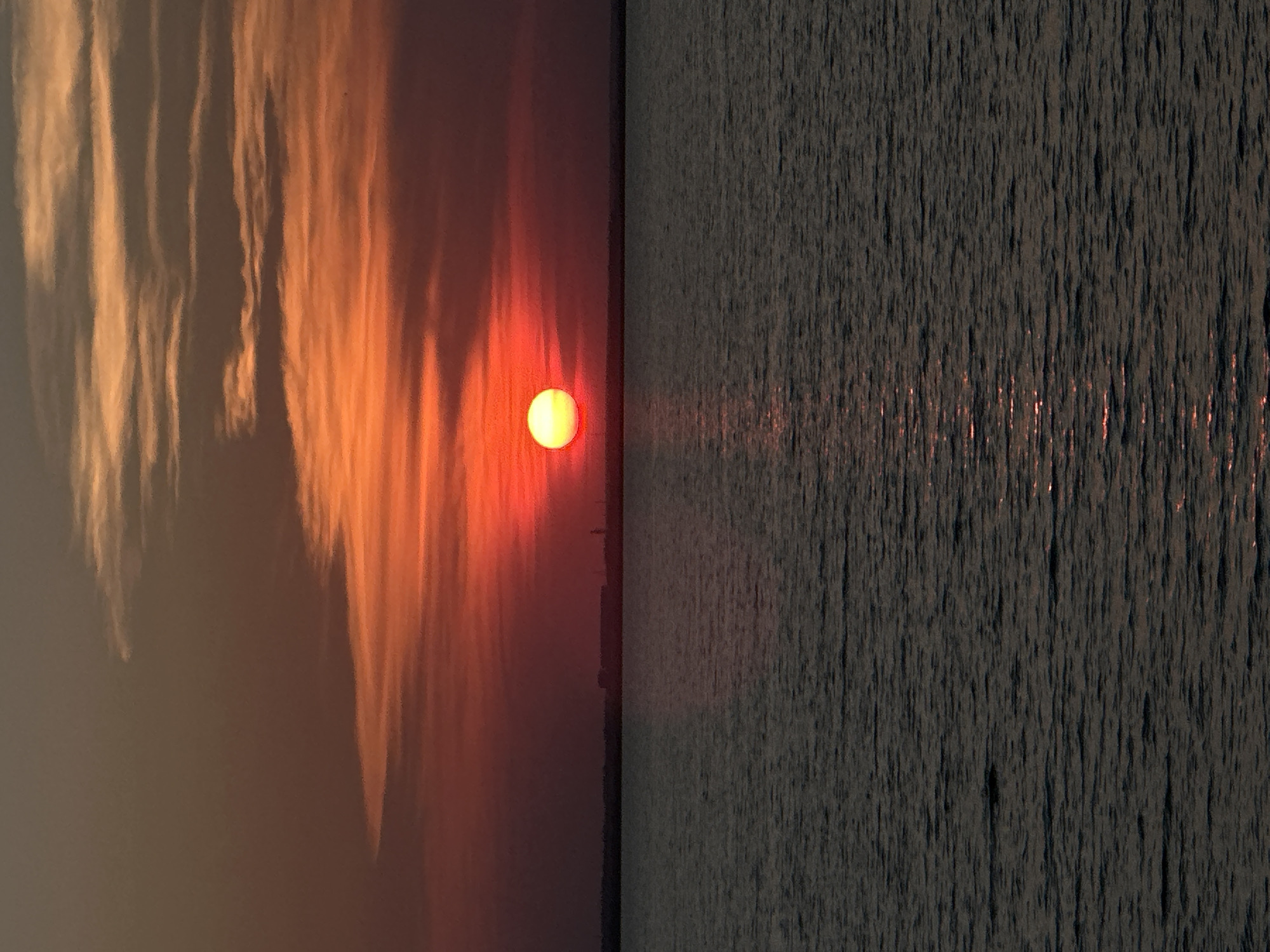 The image shows a sunset over a body of water. The sun is a bright orange orb, low in the sky, casting a warm glow on the surrounding clouds. The clouds are wispy and layered, with shades of orange, red, and purple. The water is dark and calm, reflecting the colors of the sunset. In the distance, a faint silhouette of buildings can be seen on the horizon. The overall mood is peaceful and serene. This scene is likely not in Kokshetau, Kazakhstan, as the typical landscape does not include large bodies of water that fit this description.