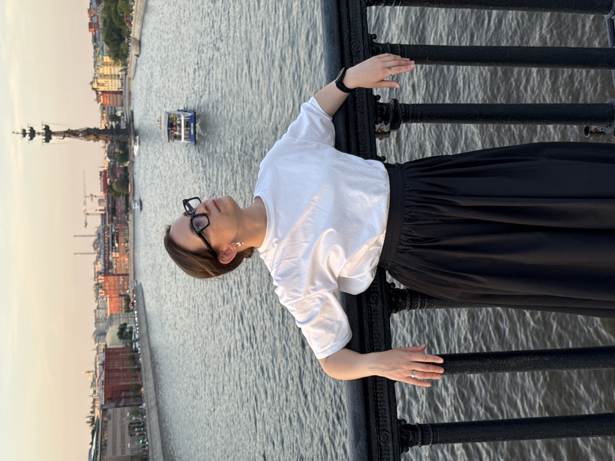 A woman stands on a bridge overlooking the Moskva River in Moscow, Russia. She wears glasses, a white t-shirt, and a black skirt. Her hands rest on the railing of the bridge. In the background, the river flows with several boats and ships present. The cityscape of Moscow is visible on the opposite bank.
