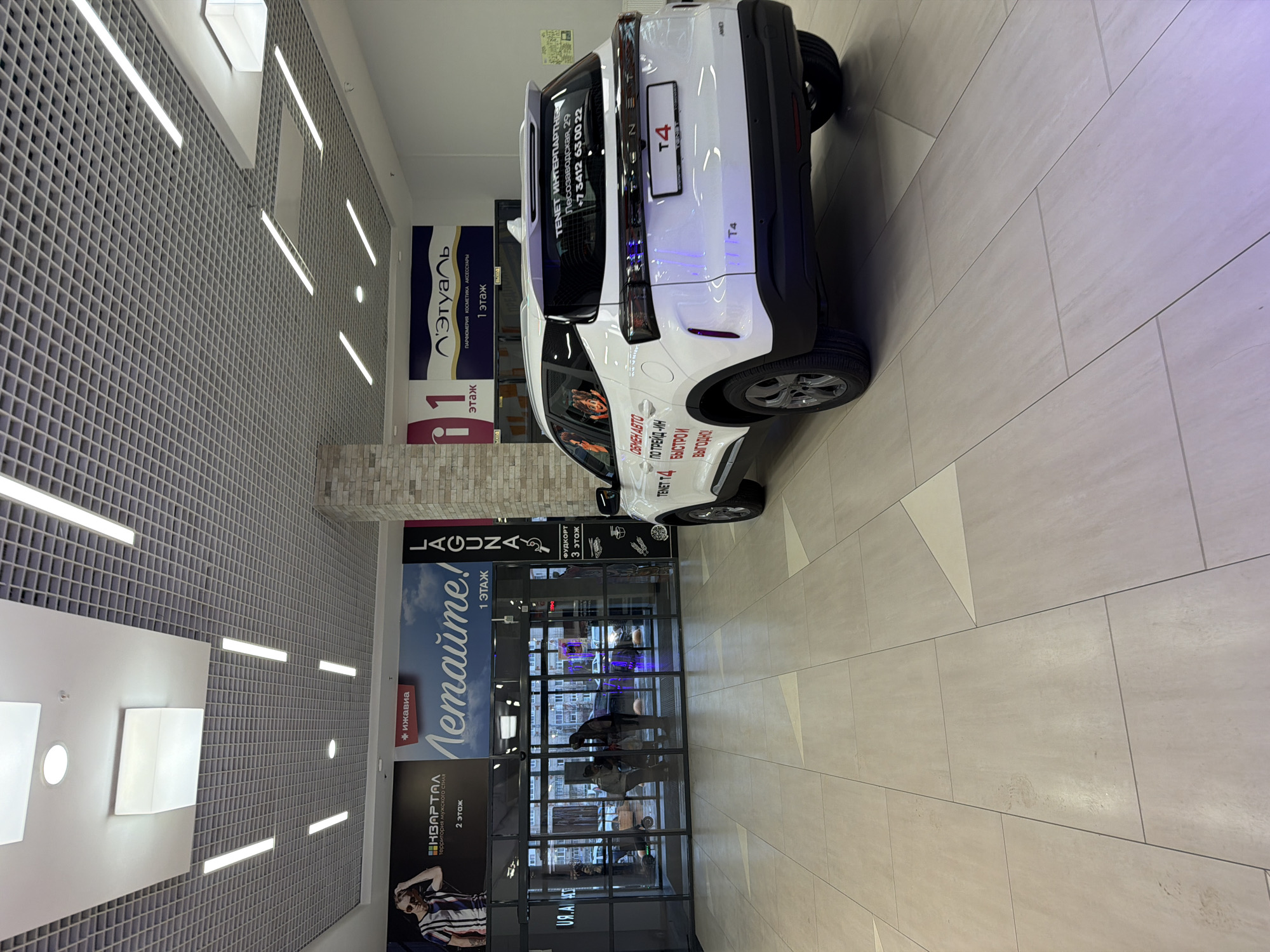 The image depicts the interior of a shopping mall in Izhevsk, Russia. A white car, labeled "T4" and bearing the "TENET ИНТЕРПАРТНЕ" (TENET INTERPARTNER) advertisement with the address "Лесозаводская, 29" (Lesozavodskaya, 29) and phone number "+7 3412 63 00 22", is parked on the mall's tiled floor. The car's advertising highlights "ОБМЕН АВТО ПО ТРЕЙД-ИН" (car trade-in) and emphasizes speed and profitability ("БЫСТРО И ВЫГОДНО!"). Behind the car is a glass-fronted retail space with several people visible inside. Mall signage indicates various businesses and levels, including "LAGUNA" (Level 1), "КВАРТАЛ" (Level 2), "Этуаль" (Level 1), and a food court on level 3. The mall's ceiling has a grid-like design with linear lighting.