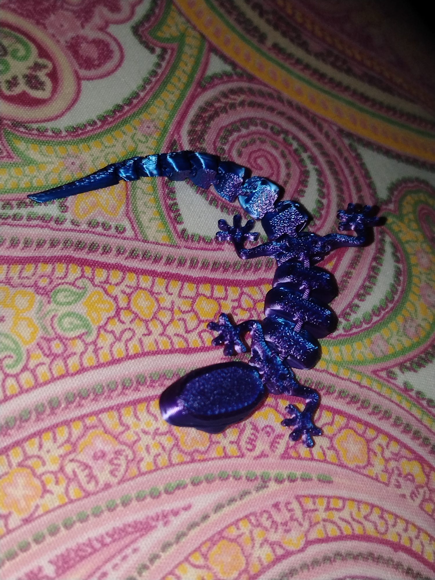 A blue articulated gecko toy is resting on a paisley patterned fabric. The paisley pattern includes shades of pink, yellow, green, and white. The gecko is primarily blue with a sparkling, iridescent sheen. No location information is present, so no reference to Izhevsk, Russia, is possible.