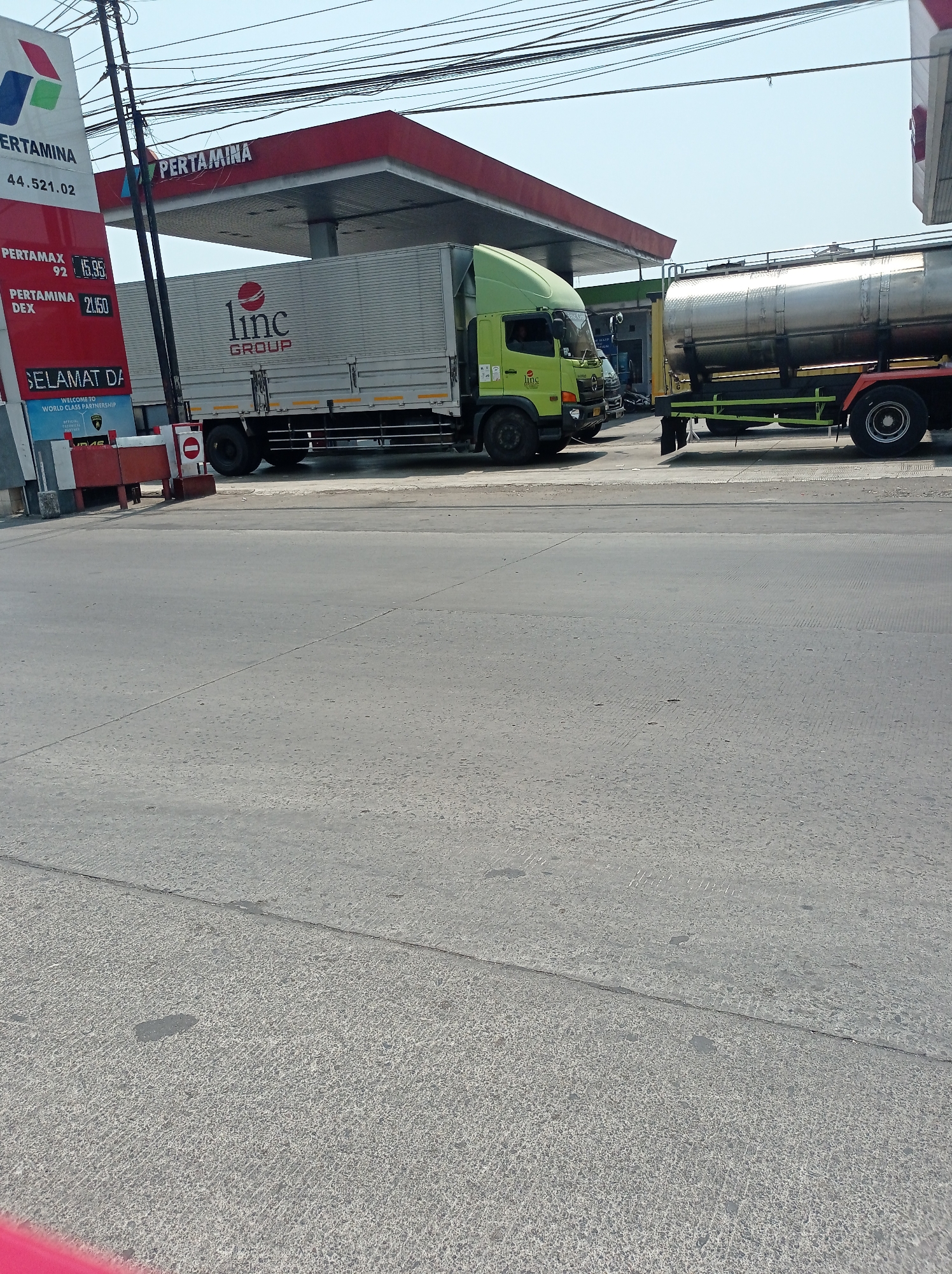 This image captures a scene at a Pertamina gas station in Dampyak, Indonesia, on a clear day. In the foreground, a grey asphalt road with visible tire marks and a thin crack runs across the frame. Behind it, a concrete surface with rough texture is visible. The middle ground is dominated by a gas station with a red roof and white pillars. Two trucks are parked at the station: a green and silver box truck with "Linc GROUP" logo on its side, and a silver tanker truck. A Pertamina signage with prices is also visible on the left side of the image. The background features a bright blue sky with thin white clouds and overhead power lines.