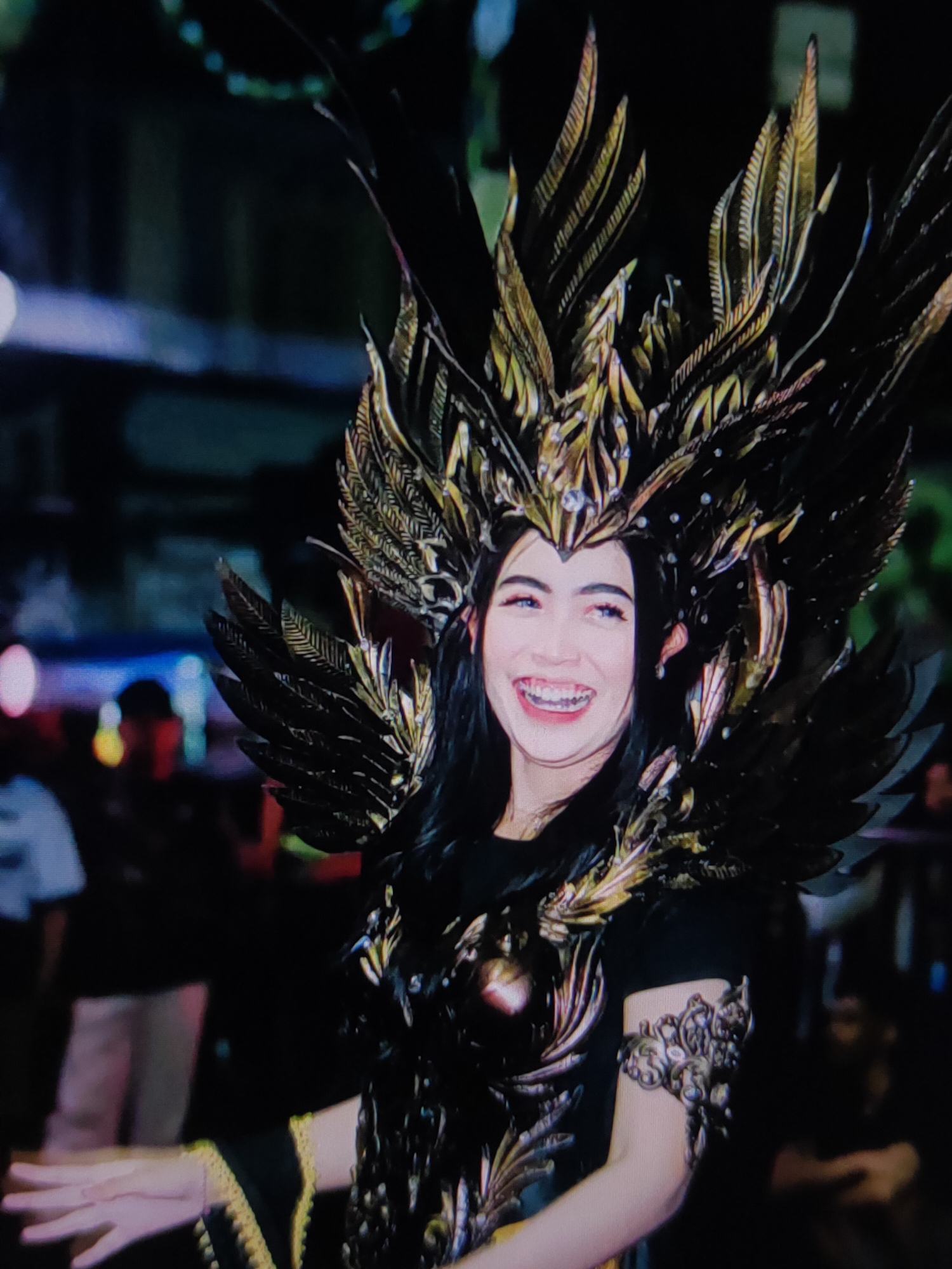 A young woman with dark hair and bangs smiles broadly at the camera, wearing an elaborate headdress and costume resembling wings and feathers. The headdress is made of black and gold materials, adorned with small, clear embellishments. She has braces on her teeth. Her costume includes a dark top and what appears to be ornate shoulder and chest armor, also in black and gold. A gold-trimmed yellow fabric is visible at the hem of her garment and on her left arm. She is wearing a decorative arm band on her right arm. In the blurred background, several people are visible, along with out-of-focus lights and dark structures, suggesting an outdoor evening event or festival. The lighting is dim, with some areas brightly lit and others in shadow.
