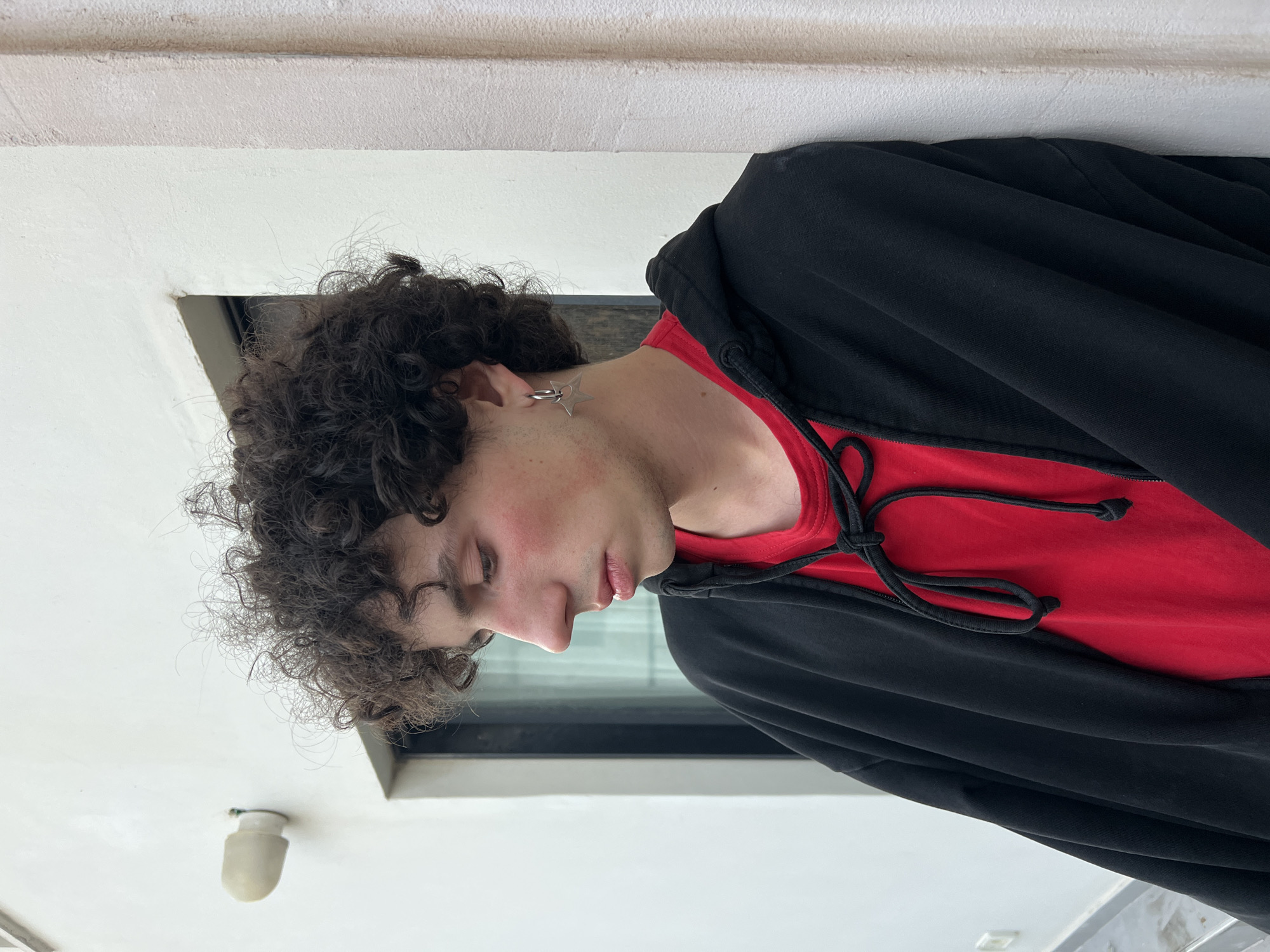 A person with curly brown hair wearing a red shirt and a black hoodie is positioned at an angle, with the head facing left. The person has an earring and appears to be in an indoor setting with white walls. A small square opening or window-like feature is visible in the background. The scene does not provide enough information to determine if it is in Tel-Aviv, Israel.