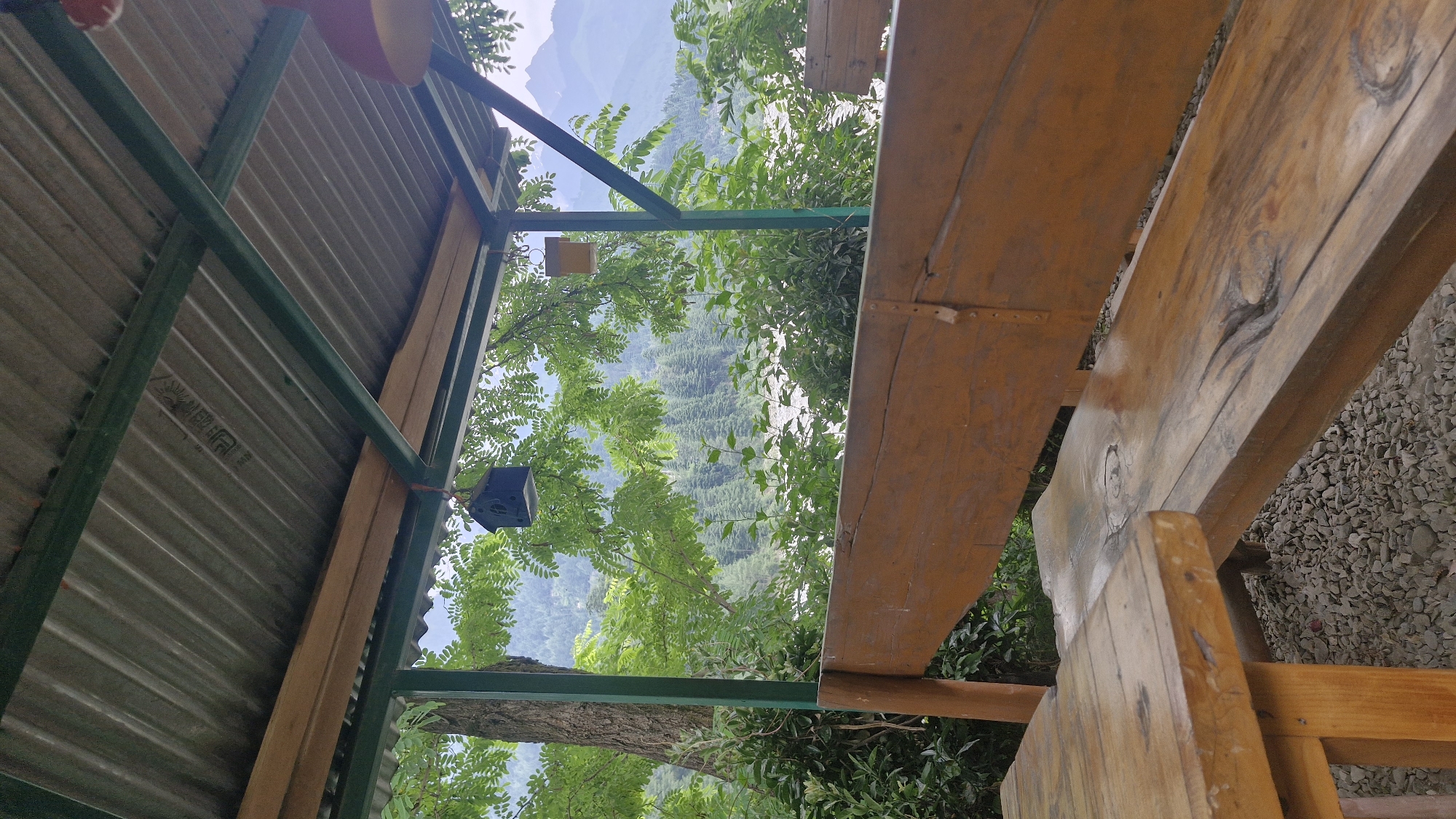 The image depicts an outdoor seating area with a rustic wooden bench and table, set against a backdrop of lush green foliage and distant mountains. The seating area appears to be covered by a corrugated metal roof supported by a green metal frame. Two decorative birdhouses, one blue and one yellow, hang from the roof. The bench is made of rough-hewn wood with visible knots and grain, suggesting a natural and perhaps aged aesthetic. The table is also wooden and has a similar rustic finish. The ground is covered in small grey pebbles. Beyond the seating area, dense green trees and shrubs are visible, with a hint of a river or body of water in the middle ground. In the distance, the soft outlines of mountains are visible under a hazy sky. The scene evokes a sense of tranquility and a connection with nature, likely a spot in or near Vashisht, India, known for its natural beauty and proximity to the Himalayas. No people are present in the image.