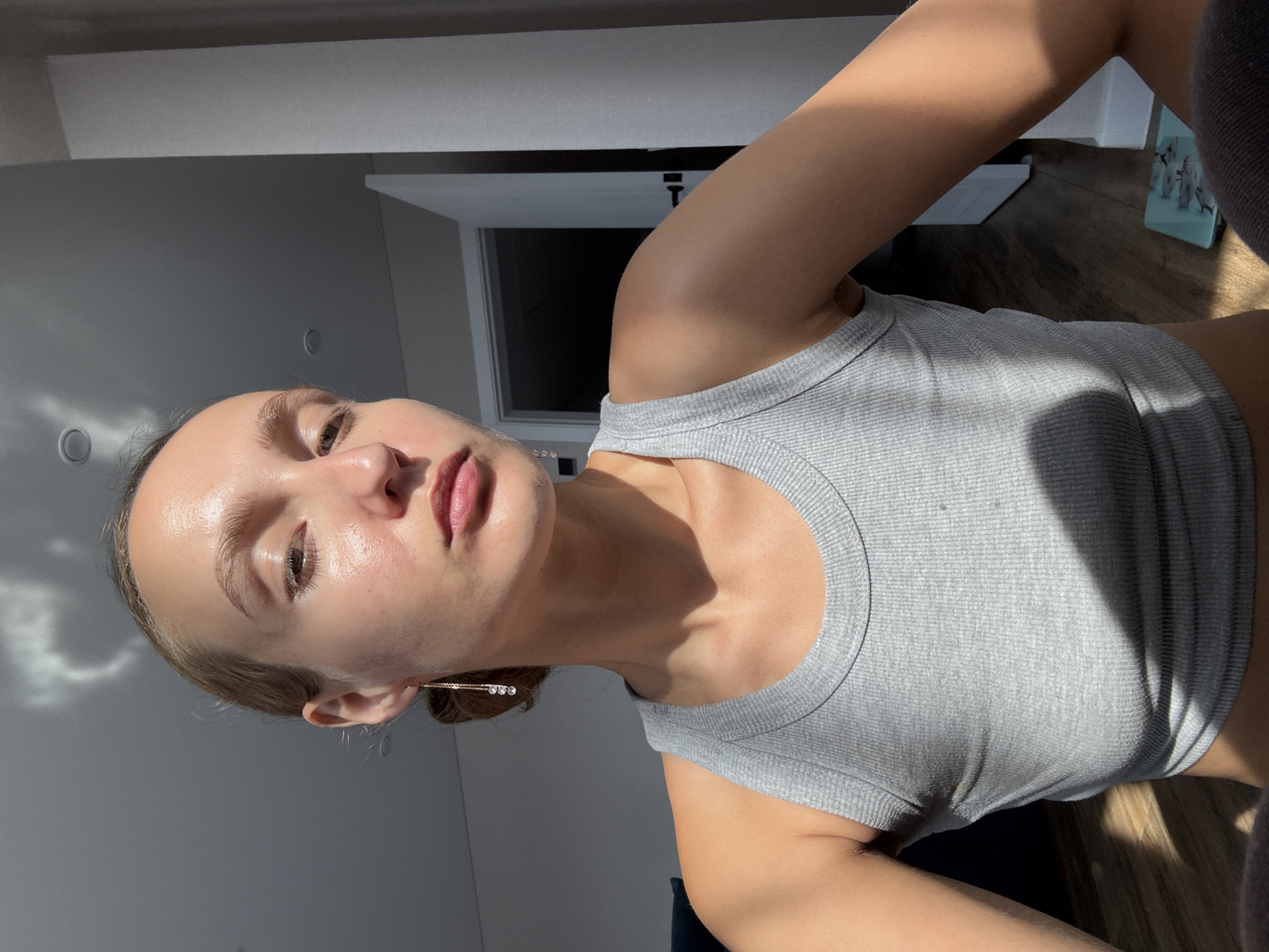 The image shows a medium shot of a young Caucasian woman with light brown hair pulled back in a bun. She is wearing a grey ribbed tank top. Her head is slightly tilted to her left, and she is looking slightly upwards and to her right. Her skin appears to be dewy and illuminated by sunlight. She has a single dangle earring with three clear beads on her left ear. The background includes a wall, a doorway, and a wooden floor. There are shadows cast by sunlight, creating a dappled effect on the woman and the floor. The woman is positioned in what appears to be an indoor setting, possibly a room in a residential building. There are no explicit indicators of the city of Подшивалово, Russia, in the image.