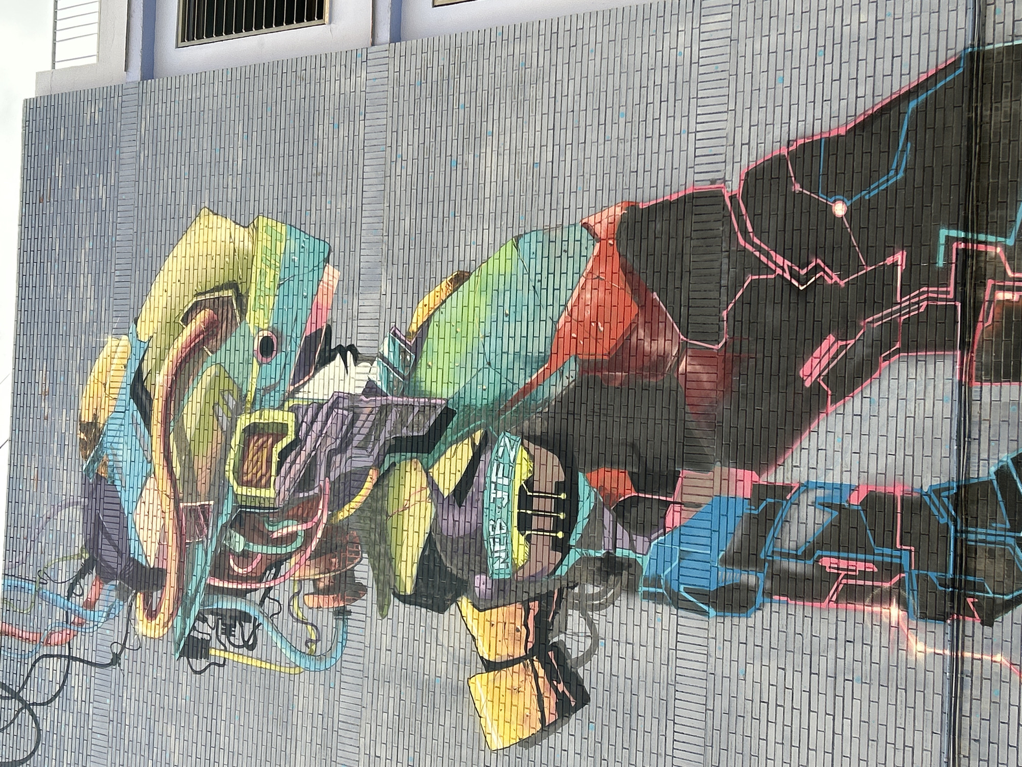 A vibrant, colorful mural depicting a stylized, robotic figure is spray-painted on a grey brick wall in Castelló De La Plana, Spain. The figure, rendered in shades of yellow, green, teal, purple, red, and pink, has a complex, mechanical head adorned with numerous tubes and wires. A prominent detail is the word "NFTBOTES" written in capital letters on a yellow panel on the figure's chest. The lower portion of the figure is rendered in darker tones with glowing neon-like lines in pink and blue, suggesting a futuristic or digital theme. The mural covers a significant portion of the brick wall, which appears to be an exterior wall of a building. To the right of the mural, a white railing and a window with horizontal slats are visible, indicating the presence of an adjacent structure. The overall scene is a striking example of urban art.
