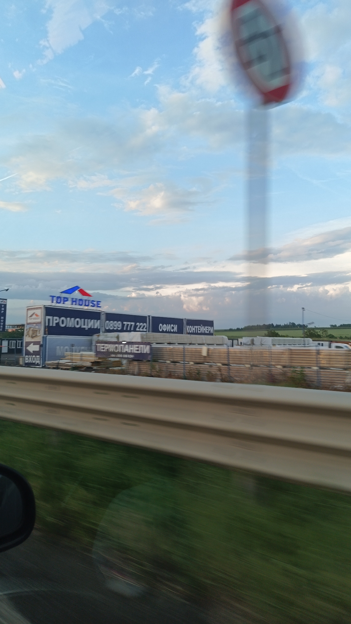 The image shows a commercial sign for "TOP HOUSE", which offers promotions, offices, and containers, with the phone number 0899 777 222. The sign also mentions "TERMOПАНЕЛИ". The image was likely taken from a moving vehicle, with a blurred road sign and vegetation in the foreground. The background shows a slightly cloudy sky. The language on the sign suggests the image was taken in Bulgaria, and the sign looks very similar to those found at Top House in Burgas, Bulgaria.