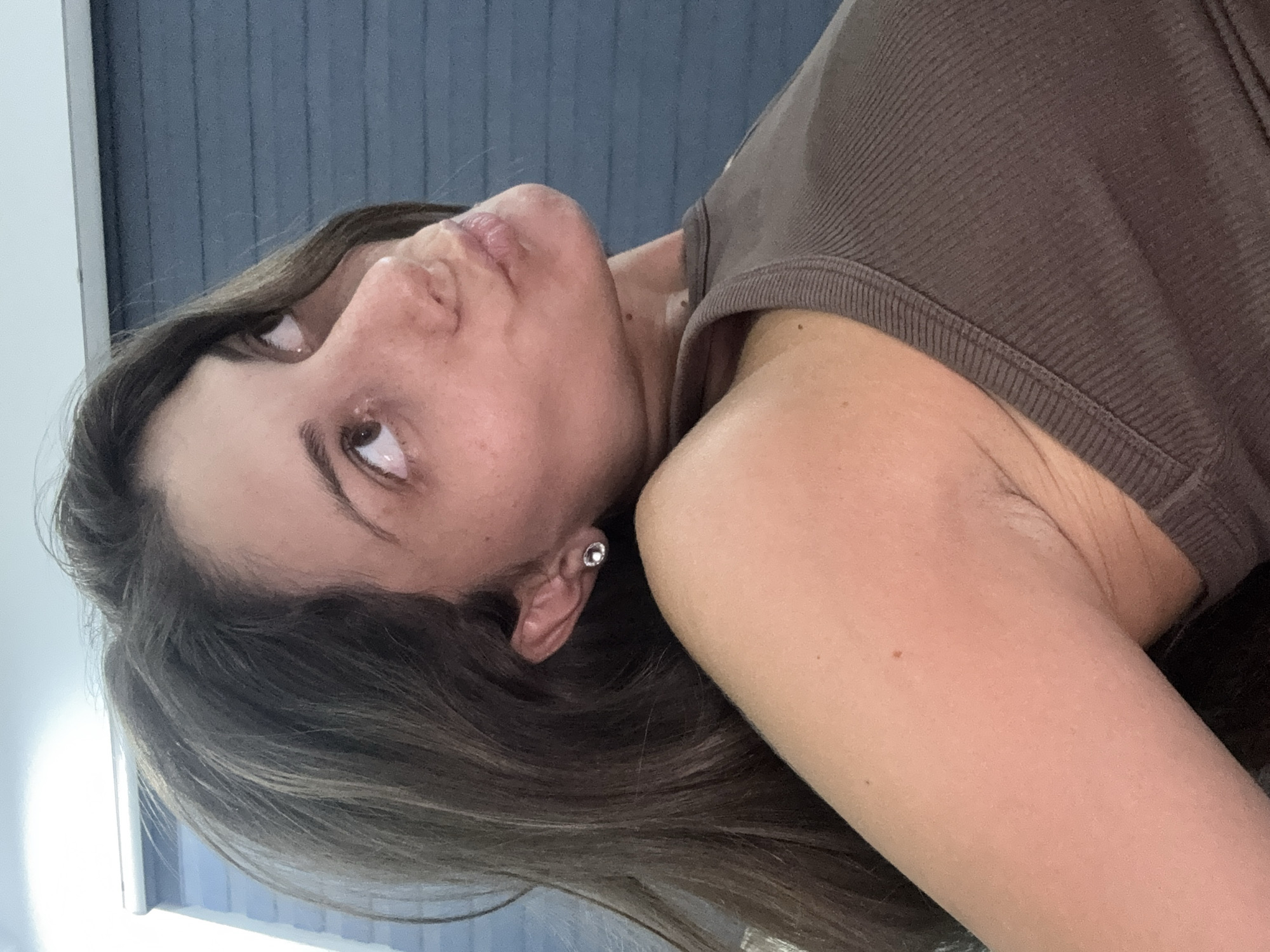 A woman with brown hair and wearing a brown tank top is positioned at an angle in the image. She has brown eyes, a small earring on her left ear, and her lips are slightly pursed. The background appears to be a textured gray wall. The location is not evident in the image.