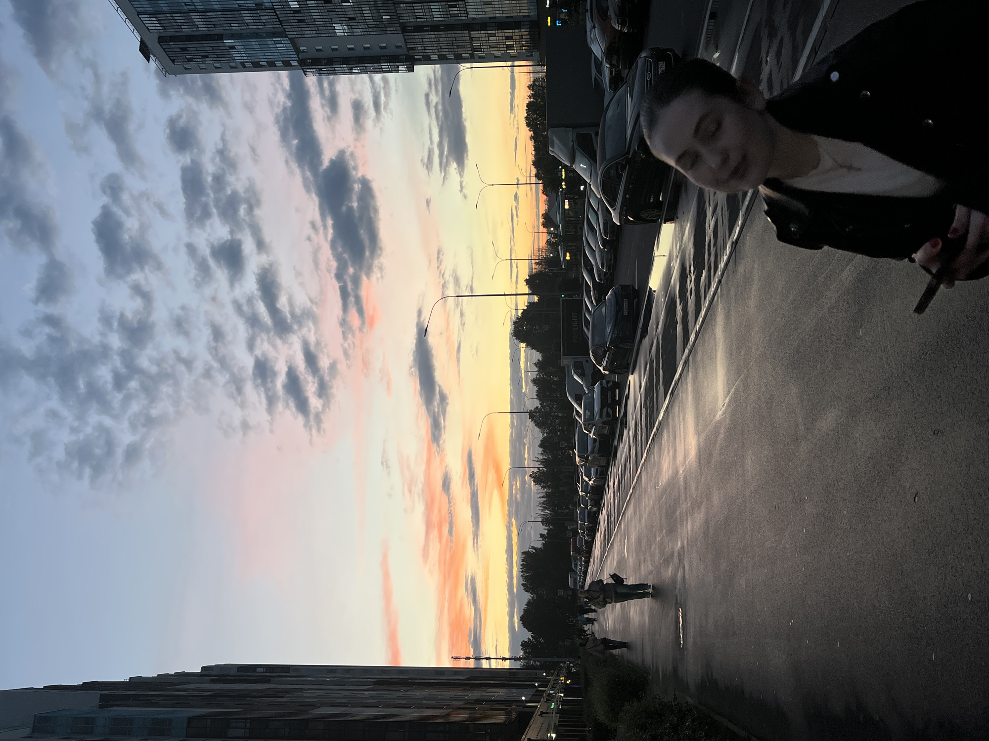 An outdoor, low-angle, full shot captures a young woman in the foreground holding a phone in her right hand, looking down at it with her eyes closed. She is wearing a black leather jacket over a white shirt and a necklace. Behind her, a wet asphalt sidewalk stretches into the distance, reflecting the warm hues of a sunset. Several people walk along the sidewalk, some heading towards the viewer and others away. A row of parked cars lines the street to the right of the sidewalk. Tall apartment buildings, one on the far left and another on the far right, dominate the background, their windows reflecting the colorful sky. The sky is filled with dramatic clouds painted in shades of pink, orange, and yellow, indicative of sunset. Streetlights dot the scene, and trees are visible in the middle ground. The overall impression is that of an urban setting in Saint Petersburg, Russia, at dusk.