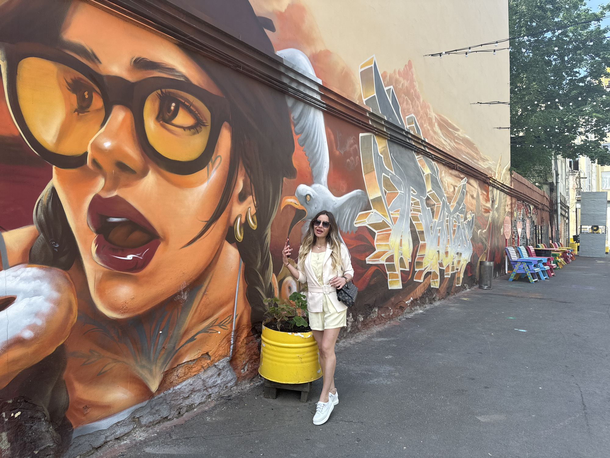 A young Caucasian woman stands in front of a vibrant graffiti mural on a textured, light-colored wall. The mural depicts a stylized portrait of a woman with large, yellow-tinted glasses and dark hair in braids. To the right of the portrait, a white bird with outstretched wings is painted, followed by abstract, geometric graffiti elements in shades of gray, orange, and yellow. The woman in the foreground is wearing a light-colored, belted blazer, matching shorts, and white platform sneakers. She has sunglasses on and is holding a smartphone in her right hand, appearing to take a selfie. A black handbag is slung over her left shoulder. In front of her and to her left, a large, bright yellow cylindrical planter holds a small green plant. The ground is paved with asphalt. Further down the alley, a row of colorful benches and a trash can are visible, alongside more buildings and trees. The scene suggests an urban outdoor setting, likely in a courtyard or alleyway. The graffiti style is reminiscent of street art commonly found in St. Petersburg, Russia.
