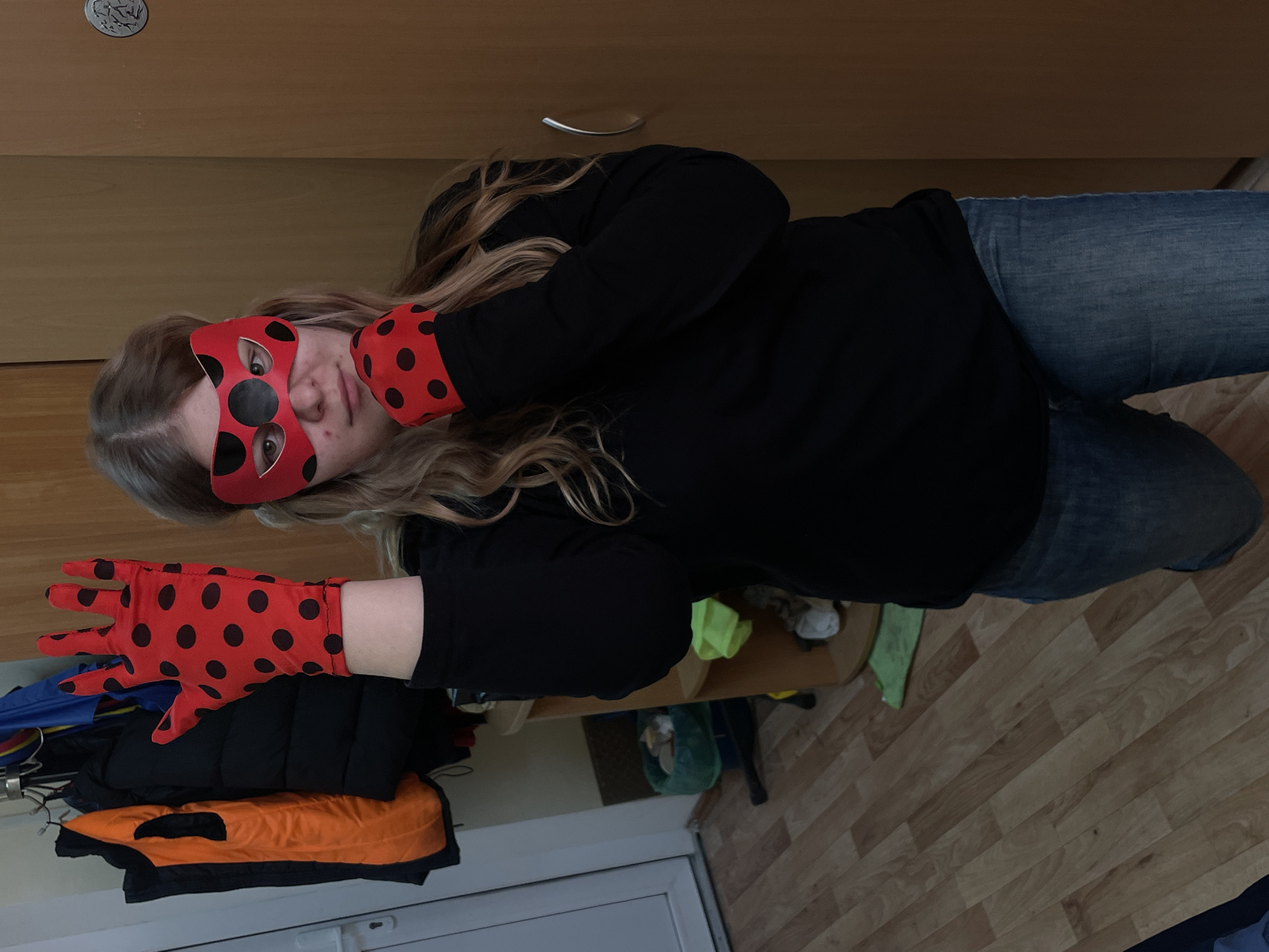 A blonde-haired individual is dressed as the character Ladybug, wearing a red mask with black spots and red gloves with black spots. They are wearing a black long-sleeve shirt and blue jeans. The location appears to be indoors, possibly a home. The room has light wood flooring, a light-colored door, and a cabinet with a silver handle. There is also a table with miscellaneous items and clothing on a rack. The location cannot be determined to be in Saint Petersburg, Russia based solely on this image.
