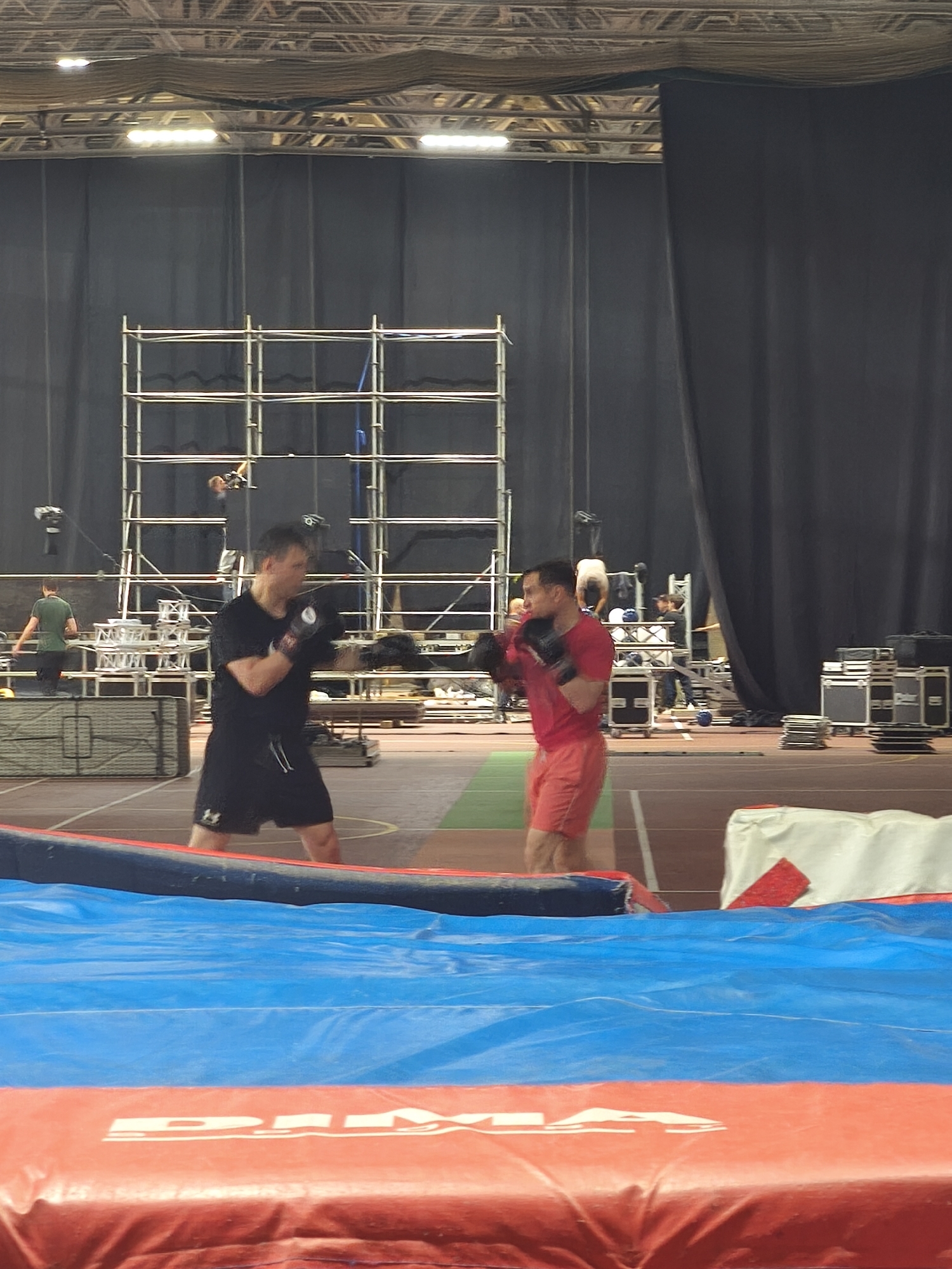 A medium, eye-level shot taken indoors at an athletic training center in Saint Petersburg, Russia. Two men are engaged in a boxing exercise. The man on the left wears black shorts and a black T-shirt and boxing gloves. The man on the right wears pink shorts and a pink T-shirt and boxing gloves.
In the background is a large, black curtain with metal scaffolding in front. Further back, additional people are working. Various equipment, including lighting and sound gear, is scattered throughout the scene. In the foreground is a blue and red padded surface. The location appears to be a gymnasium or large training facility.