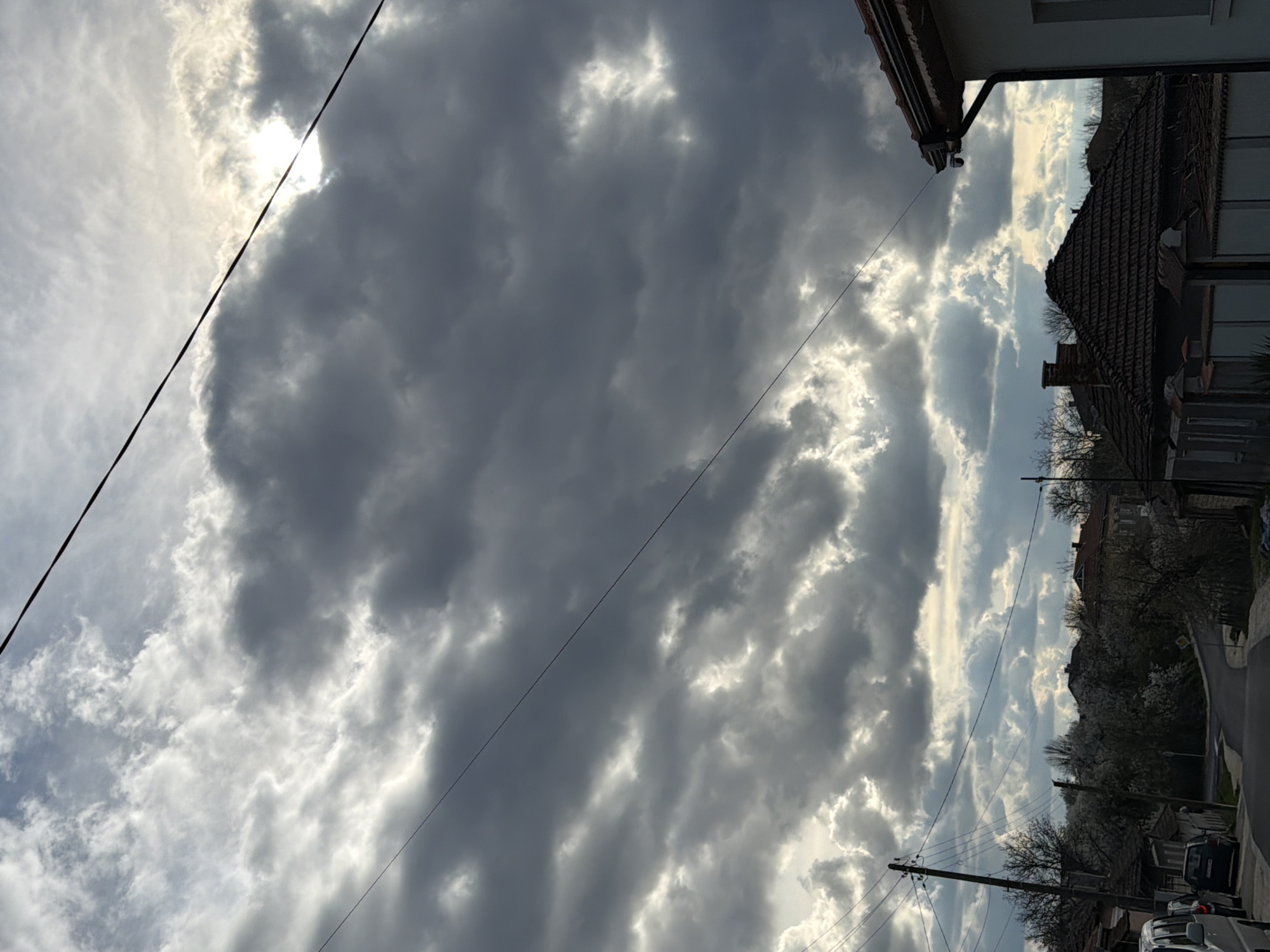 This image shows an upward view of the sky in Brestovene, Bulgaria, with dark clouds partially obscuring the sun. Utility lines are visible across the sky. Adjacent to the sky view is a partial view of buildings with tiled roofs and exterior walls, typical of residential architecture. A road and parked vehicles are also present. The sky is the predominant element, suggesting overcast weather conditions.