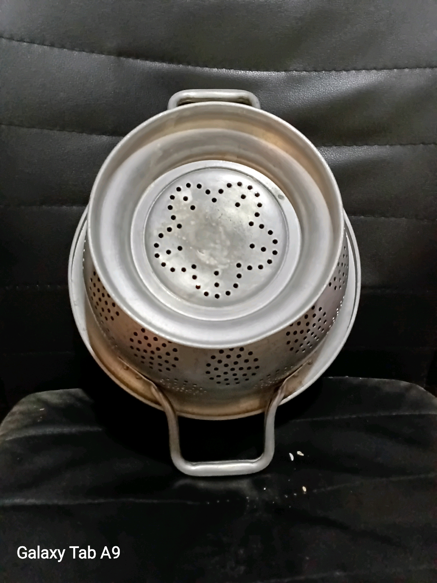 A metal colander with handles is placed on a black, textured surface. The colander has numerous small perforations throughout its body and a flat base. The surface appears to be a chair or cushion, with visible stitching lines. A watermark in the lower left corner reads "Galaxy Tab A9". The overall setting suggests an indoor environment. There is no indication of Karangan, Indonesia or any specific urban context.
