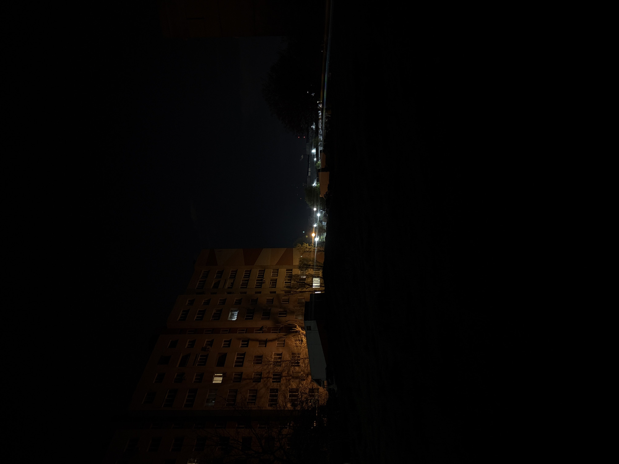 The image depicts a night scene in Sofia, Bulgaria. A multi-story building with numerous windows is illuminated with warm interior light, contrasting with the surrounding darkness. Street lights illuminate a street lined with trees and parked cars. The image is rotated 90 degrees clockwise, giving a distorted perspective. No people are visible.