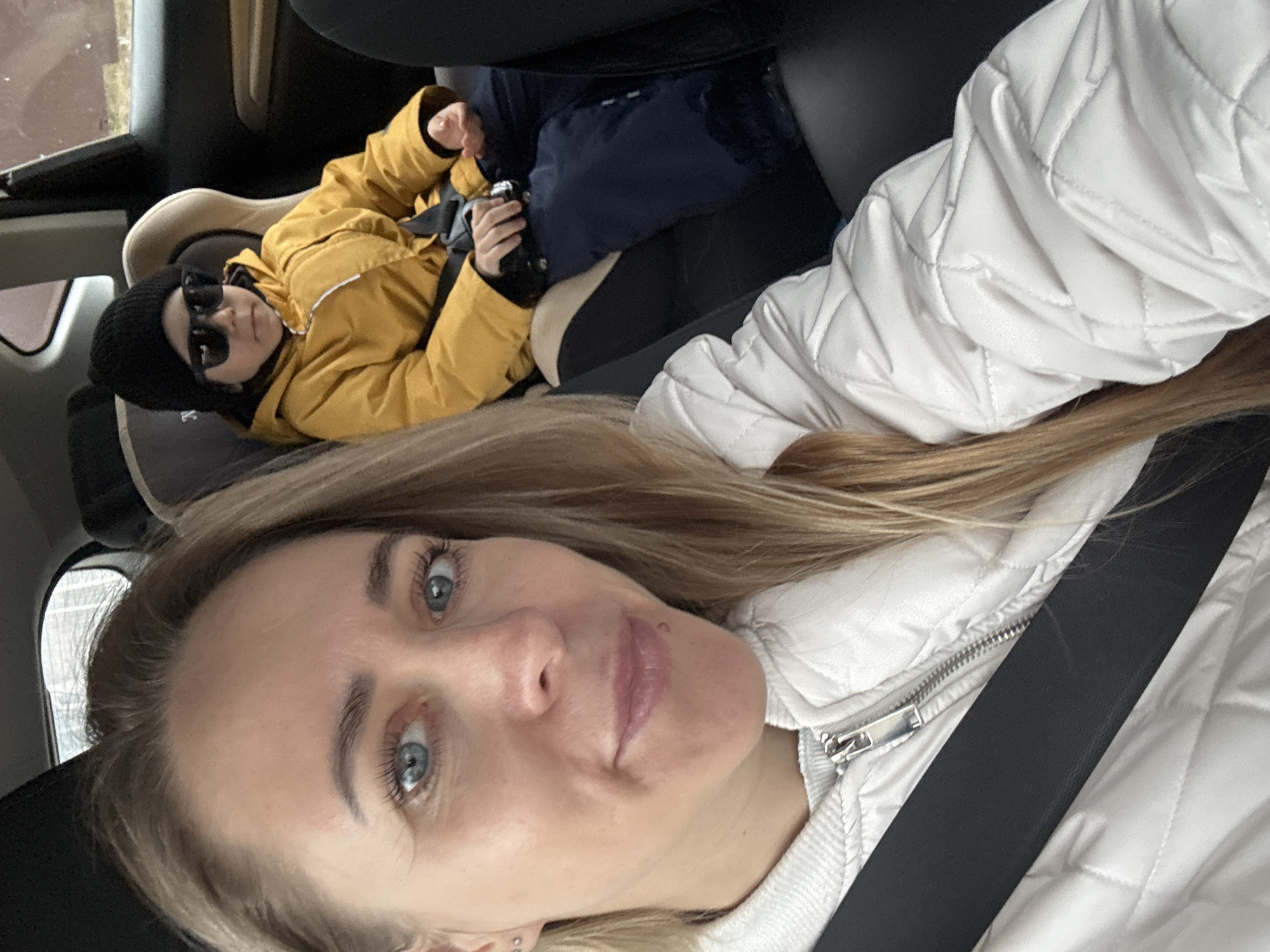 A woman with long blonde hair, wearing a white quilted jacket and a seatbelt, is taking a selfie in a vehicle. Behind her, a child in a car seat is wearing sunglasses, a black beanie, and a yellow jacket over blue pants. The child is holding a small black object in their hand. The scene appears to be inside a car in Chaikovsky, Russia, as determined by external sources and context. The environment suggests it is daytime.