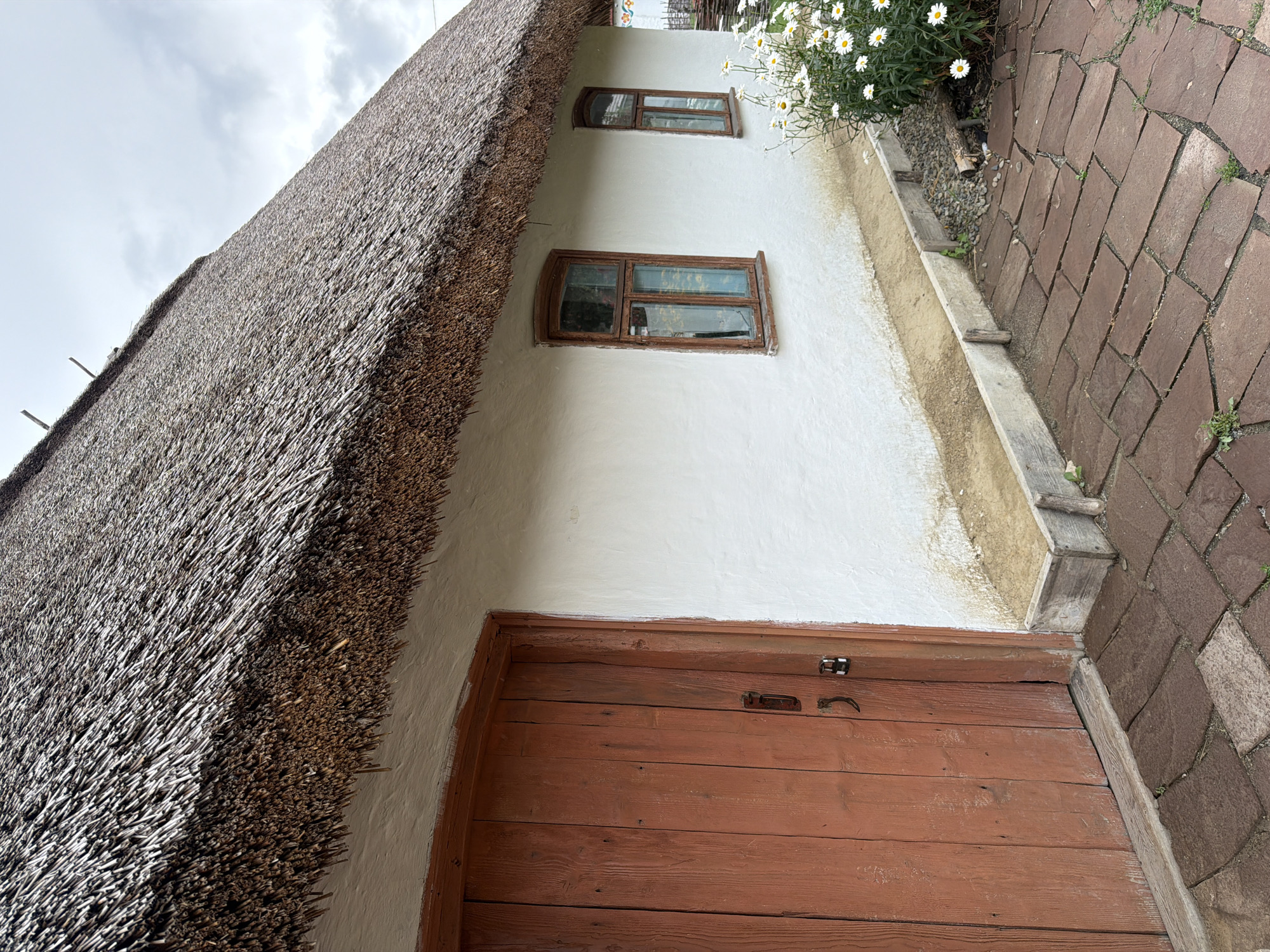 This image depicts the side of a traditional Ukrainian house in Velykyi Khodachkiv. The house features a white plastered exterior and a prominent thatched roof, characteristic of rural architecture in the region. A rustic wooden door, painted in a reddish-brown hue, is visible on the left side of the house. Two small windows with wooden frames are set into the white wall, and a bed of daisies grows near the right side of the building. The ground in front of the house is paved with reddish-brown flagstones. The sky is overcast, suggesting a cloudy day.