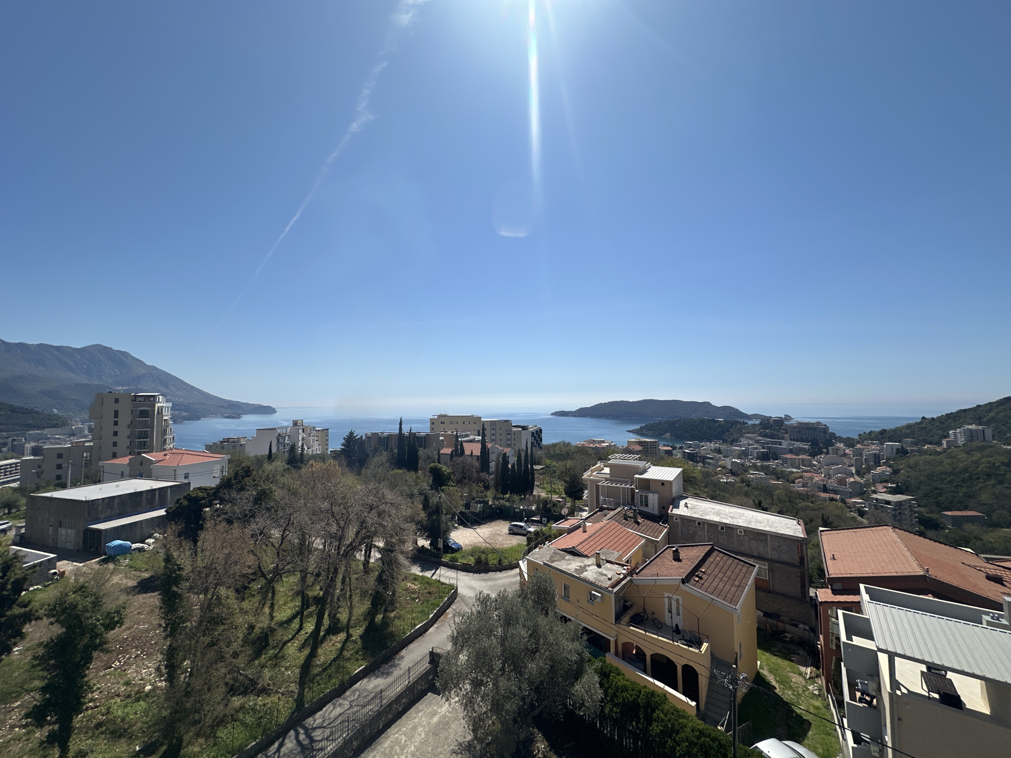 A sunny, high-angle view captures a section of Boreti, Montenegro. In the foreground, residential buildings with orange and grey roofs are visible, alongside streets and landscaping. Beyond these structures, the Adriatic Sea stretches toward the horizon, framed by mountains on either side. The sky is clear blue with a contrail and bright sunshine.