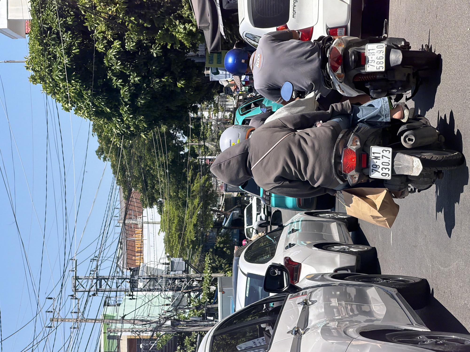 A bright, sunny day in Bac Nha Trang Ward, Vietnam, is depicted in this street-level view of traffic congestion. Multiple vehicles, including cars and motorcycles, are lined up on the asphalt road, indicating a slowdown. Two individuals are riding a scooter in the foreground, with the person in the back wearing a brown hooded jacket and blue jeans, and the person in front wearing a grey helmet and a light-colored garment. The scooter has a brown paper bag tucked behind the driver. Another scooter is visible behind them, with a rider wearing a grey shirt and a blue helmet. Cars, including a white sedan and a white SUV, are in adjacent lanes. Overhead, a dense network of electrical wires crisscrosses the sky, supported by numerous utility poles. Trees with lush green foliage line the street, and buildings with signage, including what appears to be a hotel sign with Vietnamese text, are visible in the background. The overall scene is characteristic of a busy urban environment during daylight hours.