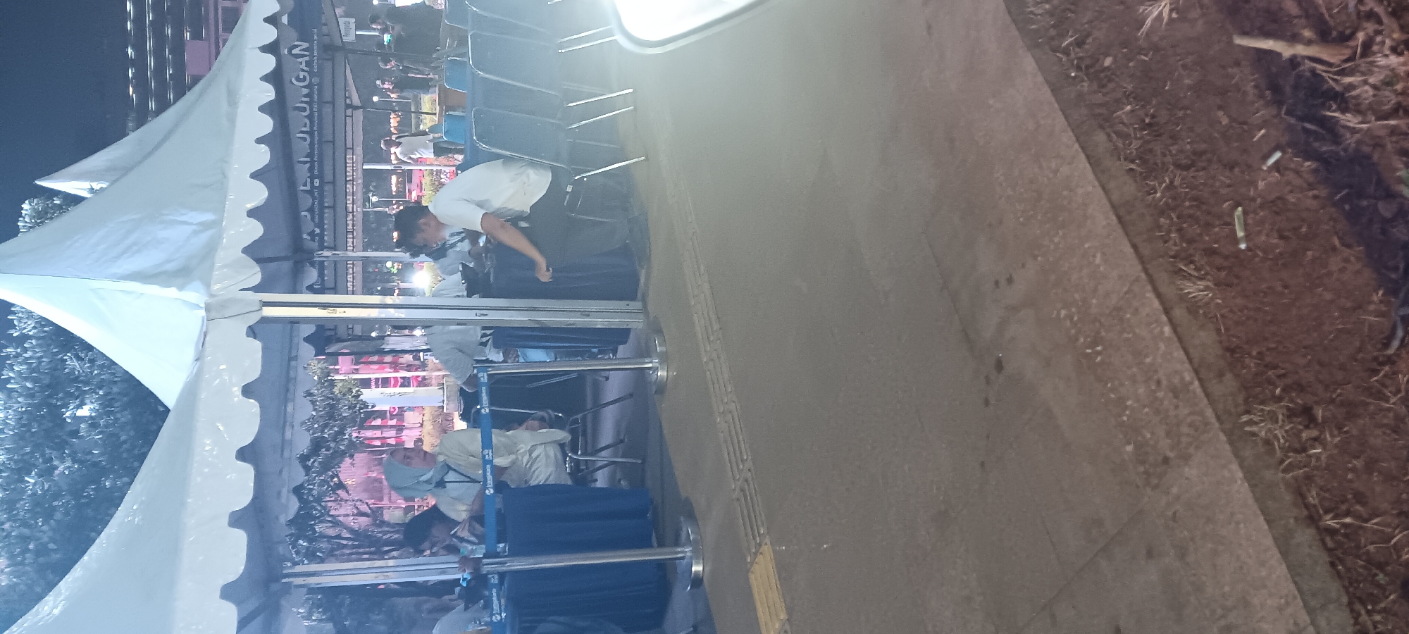 The image depicts an outdoor scene at night, possibly a registration or information booth area, under white tents. In the foreground, a paved walkway with tactile paving is visible. Behind this, several blue-draped tables with silver stanchions and blue ropes delineate a queue or waiting area. People are seated at these tables or in chairs nearby. One woman wearing a light blue hijab and a light-colored outfit is seated at a table, looking to her left. To her right, a man in a white shirt and dark pants is seated, facing away from the camera and looking to his left. Several other individuals are present in the background, some seated and others standing. The environment suggests a public event or service setup. A banner with Indonesian text, including "JISUNGAN," is visible behind the tents. The lighting is artificial, casting a dim glow on the scene. The overall impression is of an organized gathering, possibly a government service point or a registration desk for an event.