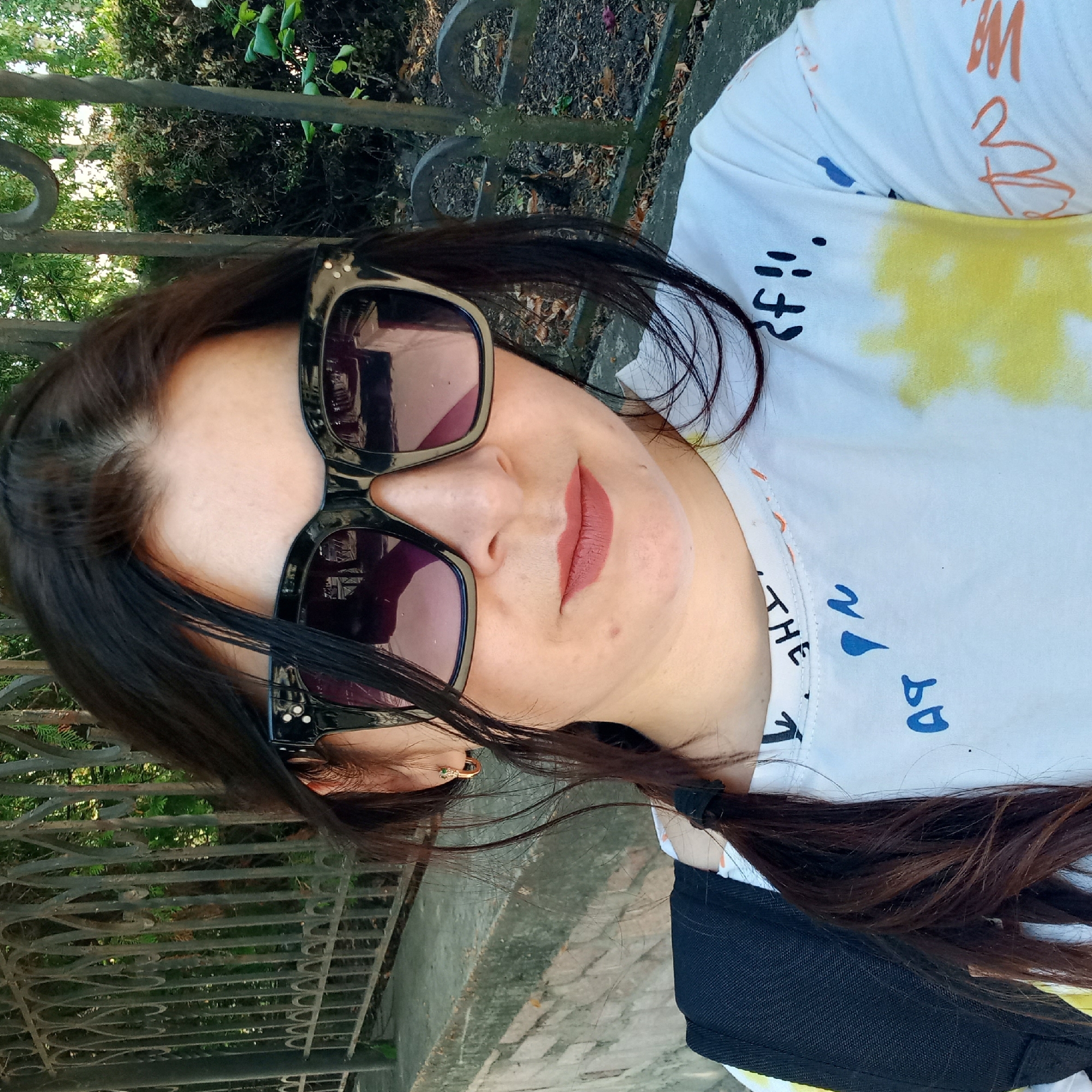 The image is a close-up, selfie-style photograph of a woman wearing sunglasses and a white t-shirt with colorful graphics and text. She has dark hair pulled into a braid. She is outdoors, with a metal fence and greenery in the background. Based on the provided context, the location is Kropyvnytskyi, Ukraine. The scene is daytime, and the lighting suggests an outdoor setting.