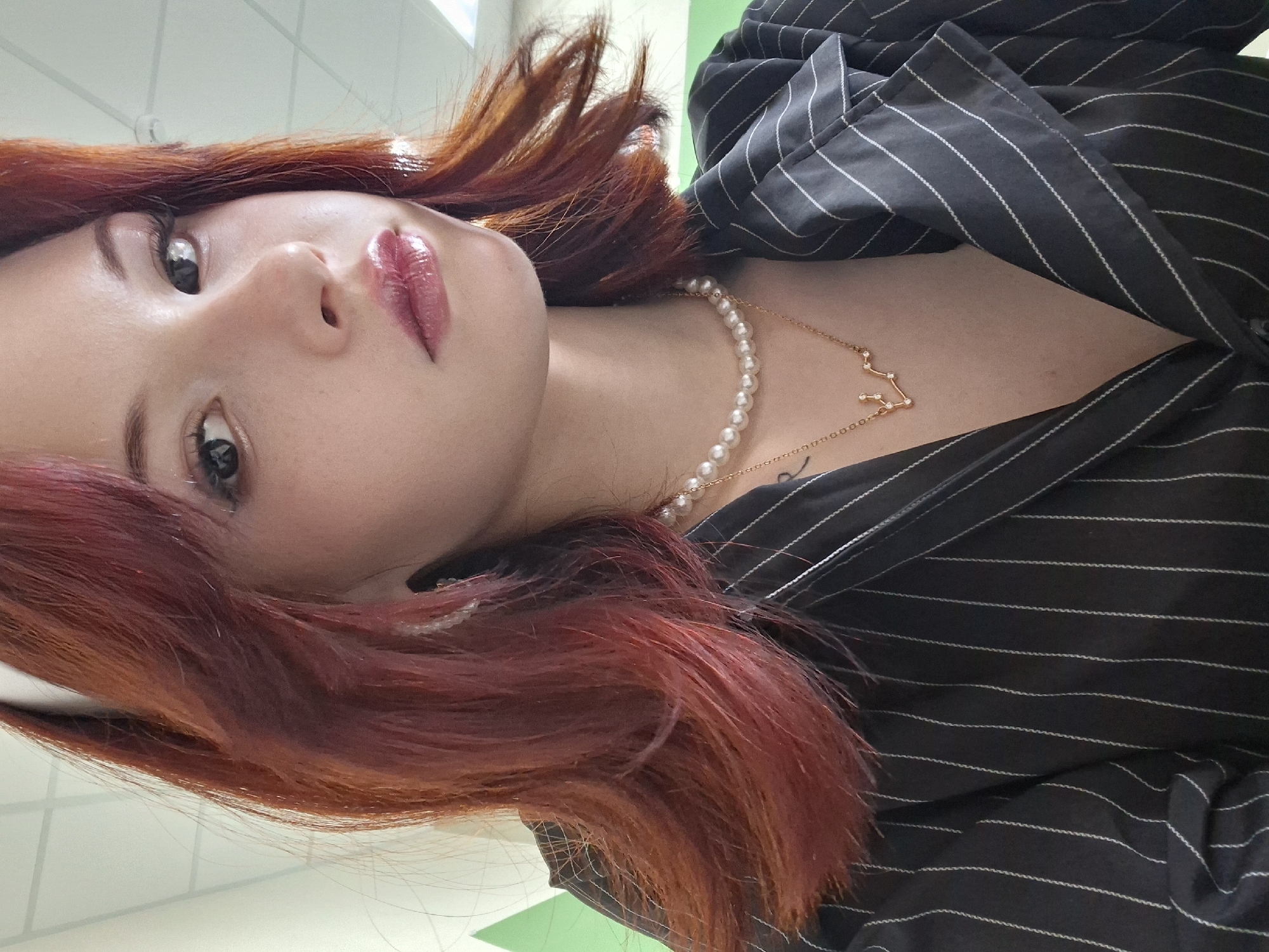A close-up, rotated portrait of a woman with red hair, wearing a black striped blazer, a pearl necklace, and a gold necklace with a constellation pendant. The background shows a glimpse of an interior with a white and green wall. There is no indication of Chaikovsky, Russia, or any specific location.