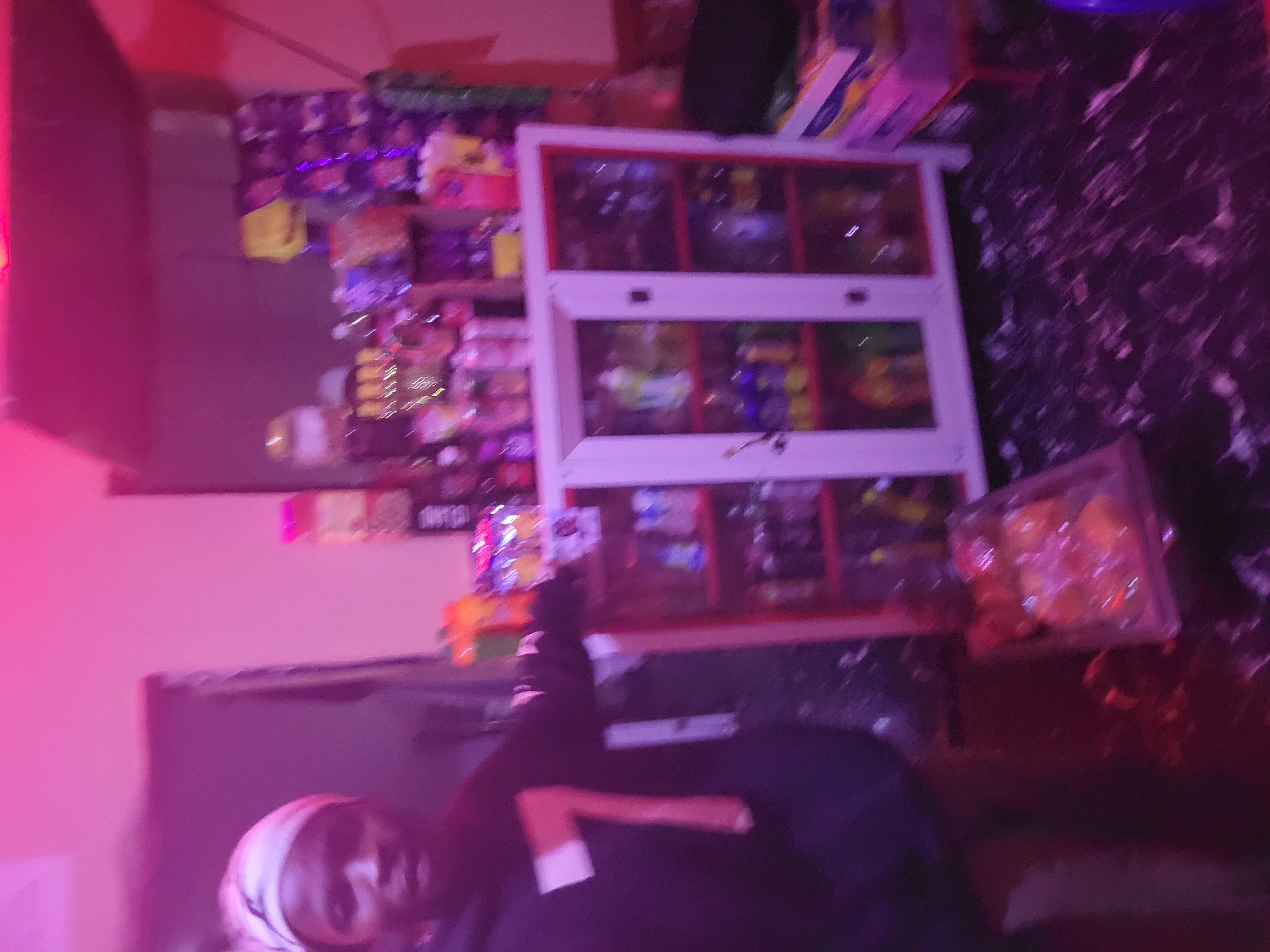 A person wearing a white headscarf and a dark hoodie with a large "7" graphic on it stands in what appears to be a small store or stall. The store is dimly lit, with bright pink and purple lighting casting an atmospheric glow. Behind the person is a white cabinet with glass doors, filled with various bottles and products. Shelves above the cabinet are also stocked with more items, including what look like packaged snacks and drinks. The floor is dark and patterned, possibly with marble or a similar material. A small, clear plastic container with some yellow items is visible on the floor to the right. The overall impression is of a modest retail space, possibly a convenience store or a small shop, with an inventory of beverages and packaged goods. The location is identified as Durumi, Nigeria.